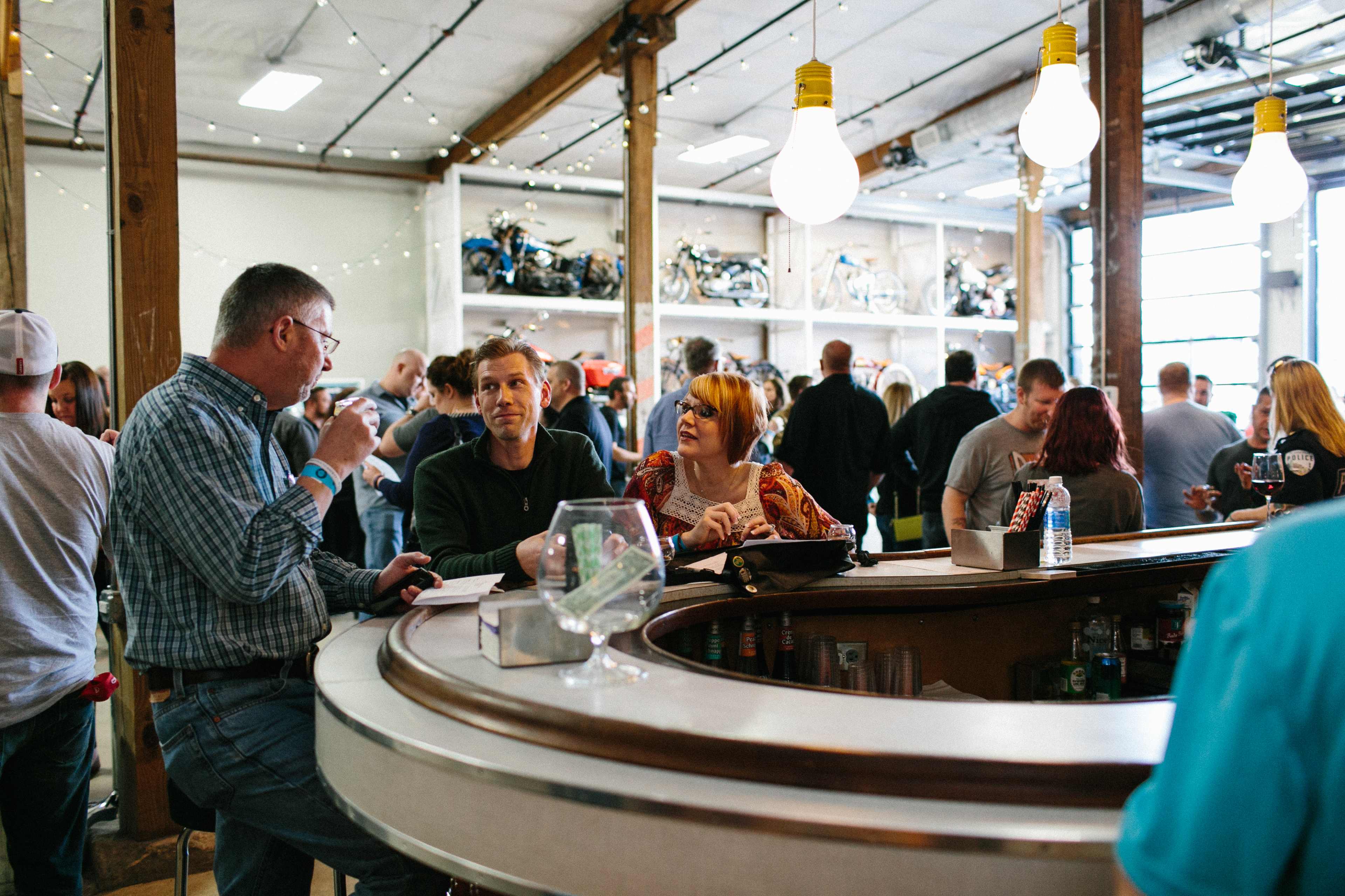 A group of people converse at a circular bar in a busy venue filled with motorcycles on display.