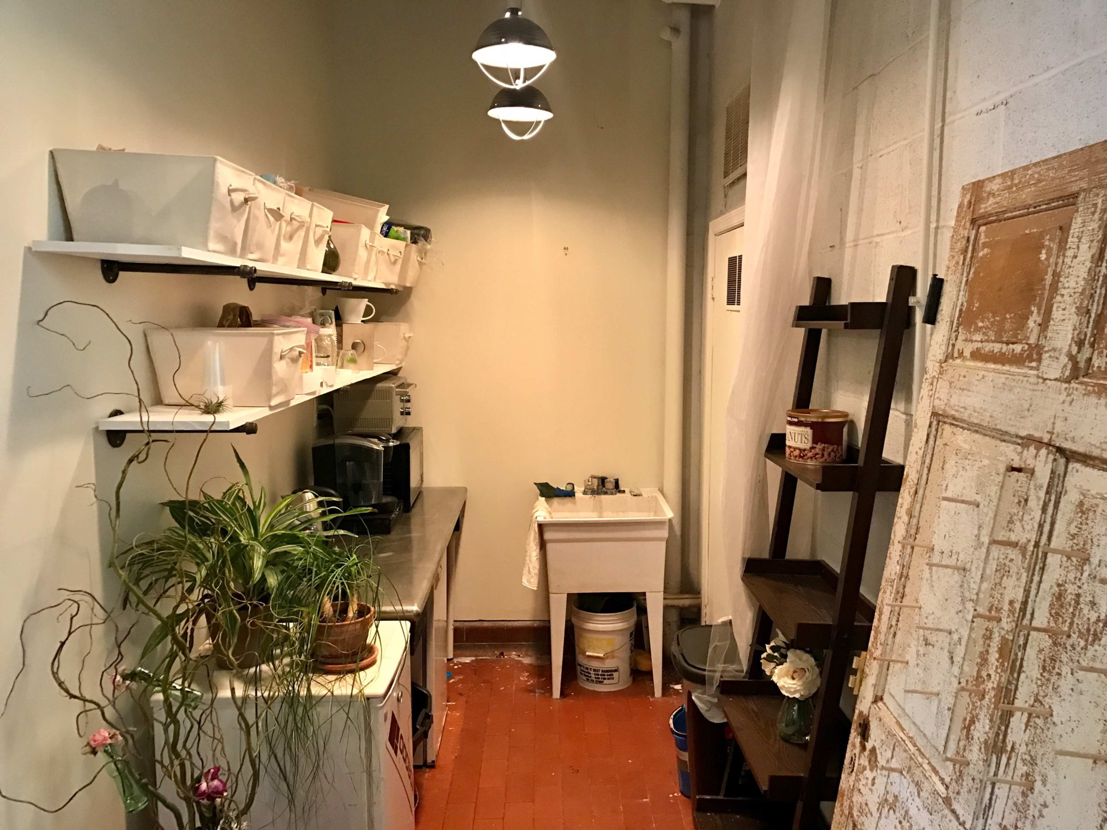The image shows a small kitchen area with a stainless steel countertop, shelves containing storage boxes, a sink, and a wooden door leaning against the wall.