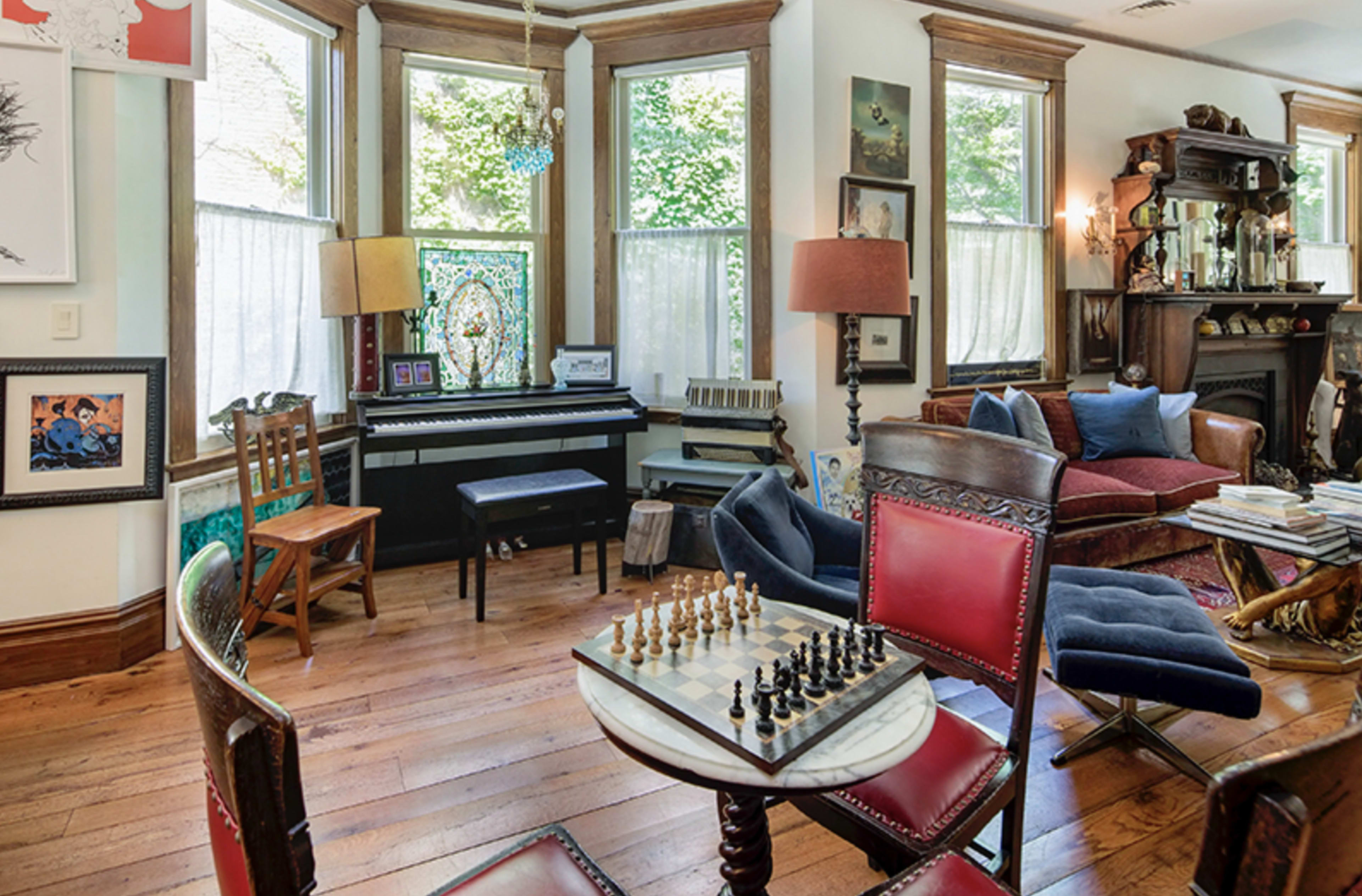 A living room features a chess set on a round table, a piano beside large windows, and various pieces of furniture arranged around the space.