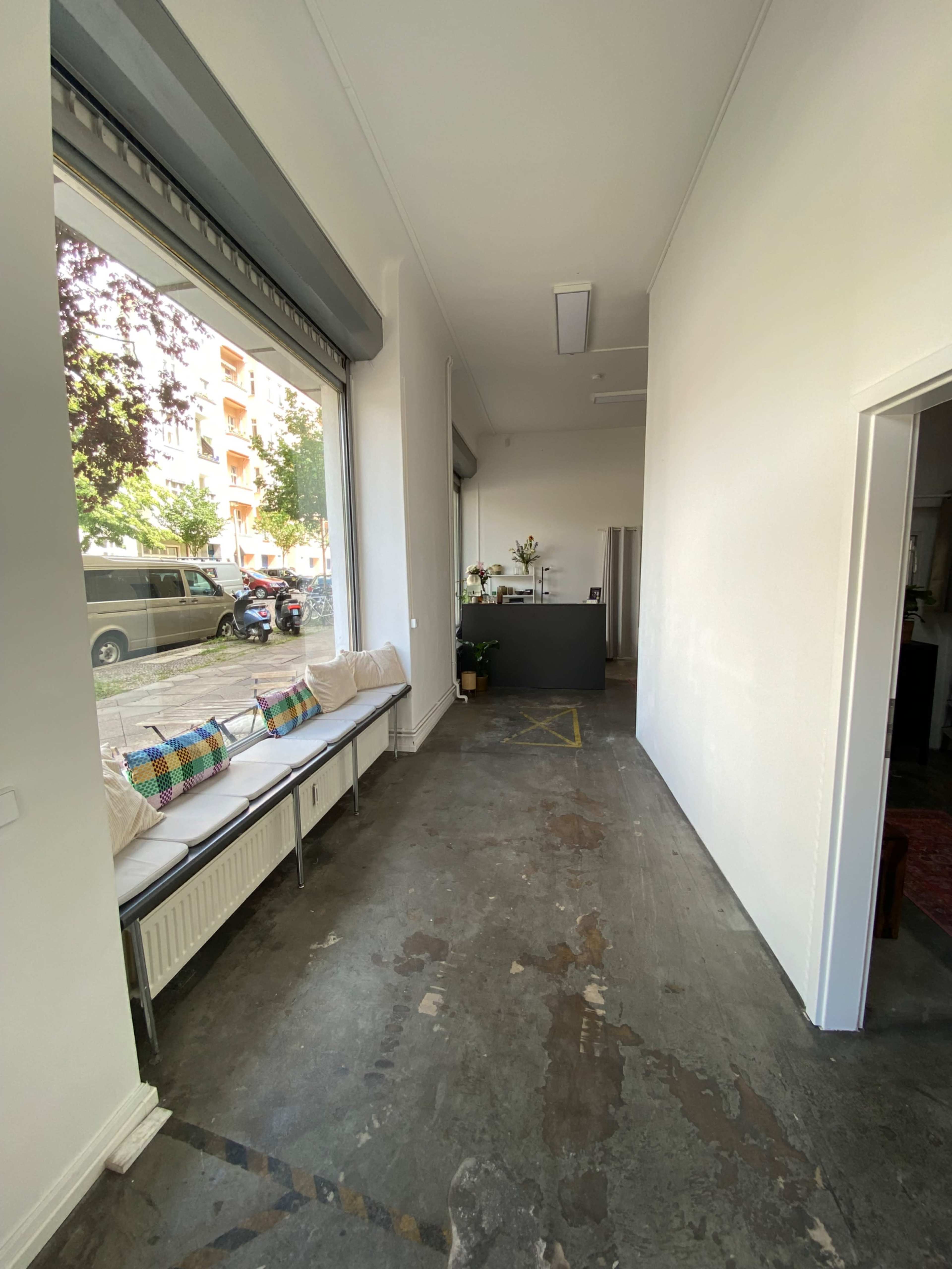 The image shows a corridor with large windows on one side, a seating area with cushions, and a view of the street outside, while a counter is positioned at the far end.
