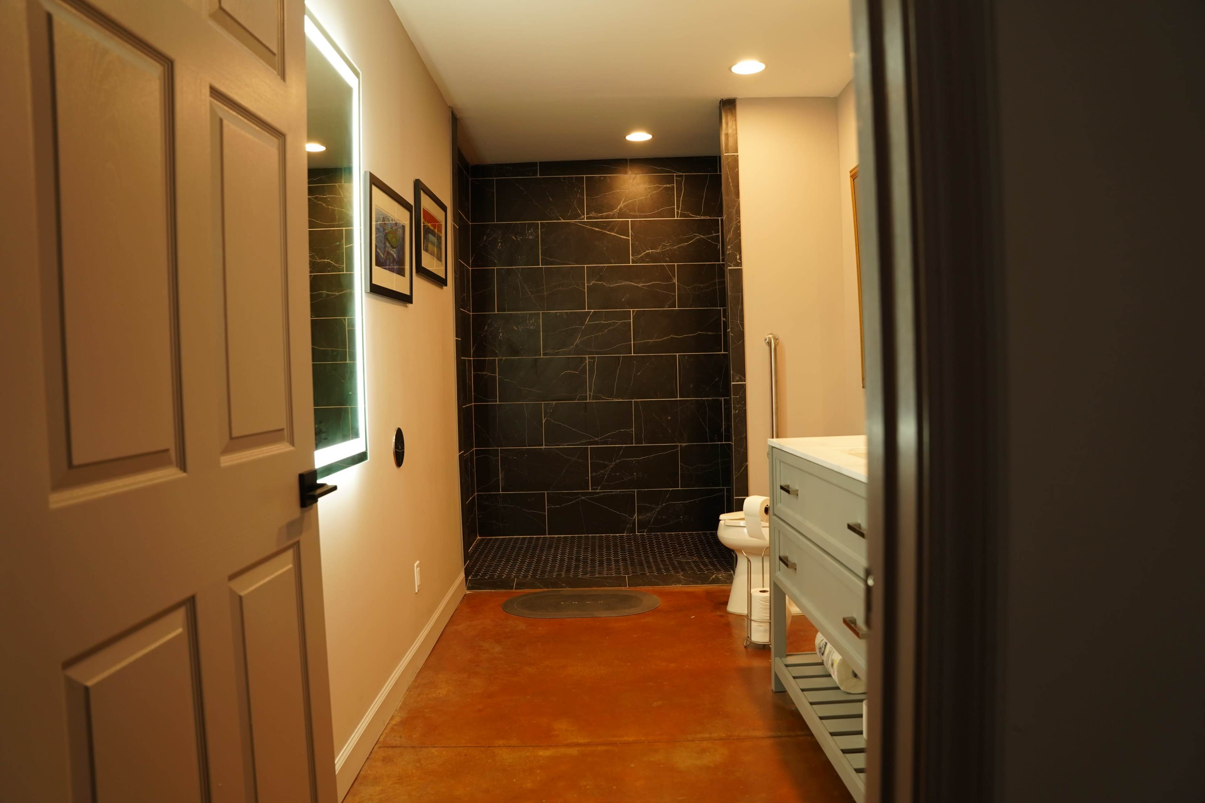 A bathroom featuring a walk-in shower, a toilet, and a sink cabinet with towels on a shelf, illuminated by overhead lights.