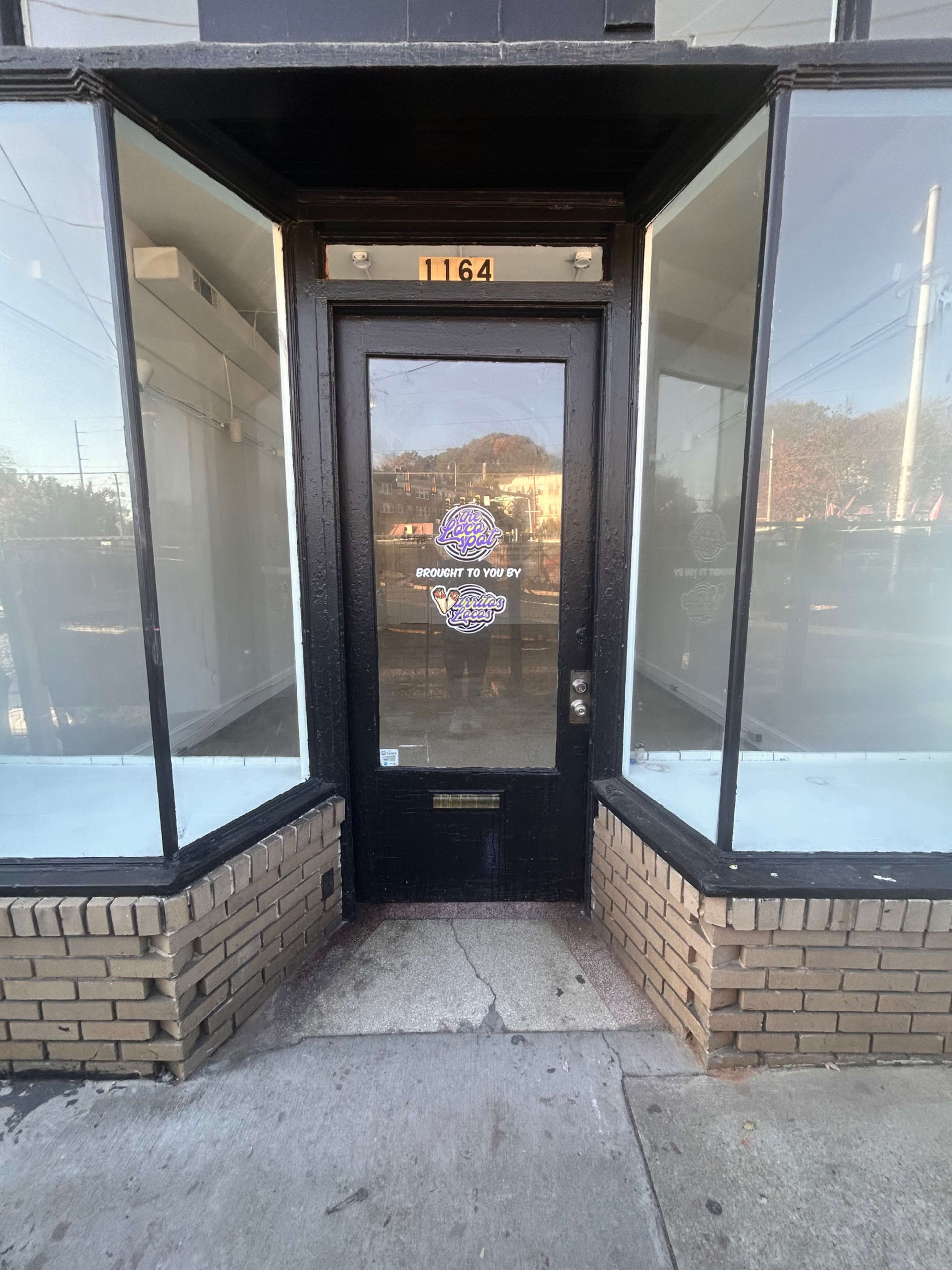 The image shows a black door with a glass storefront on either side, situated at the corner of a building.
