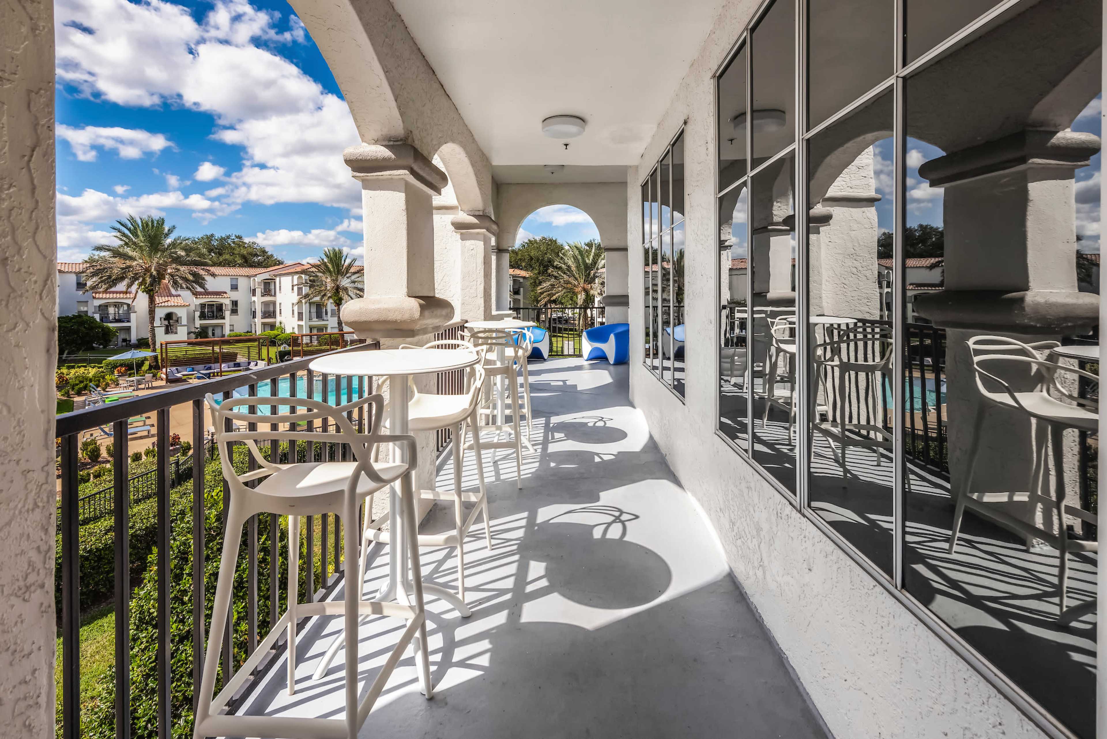 The image shows a balcony with modern bar stools overlooking a pool area surrounded by palm trees and several buildings.