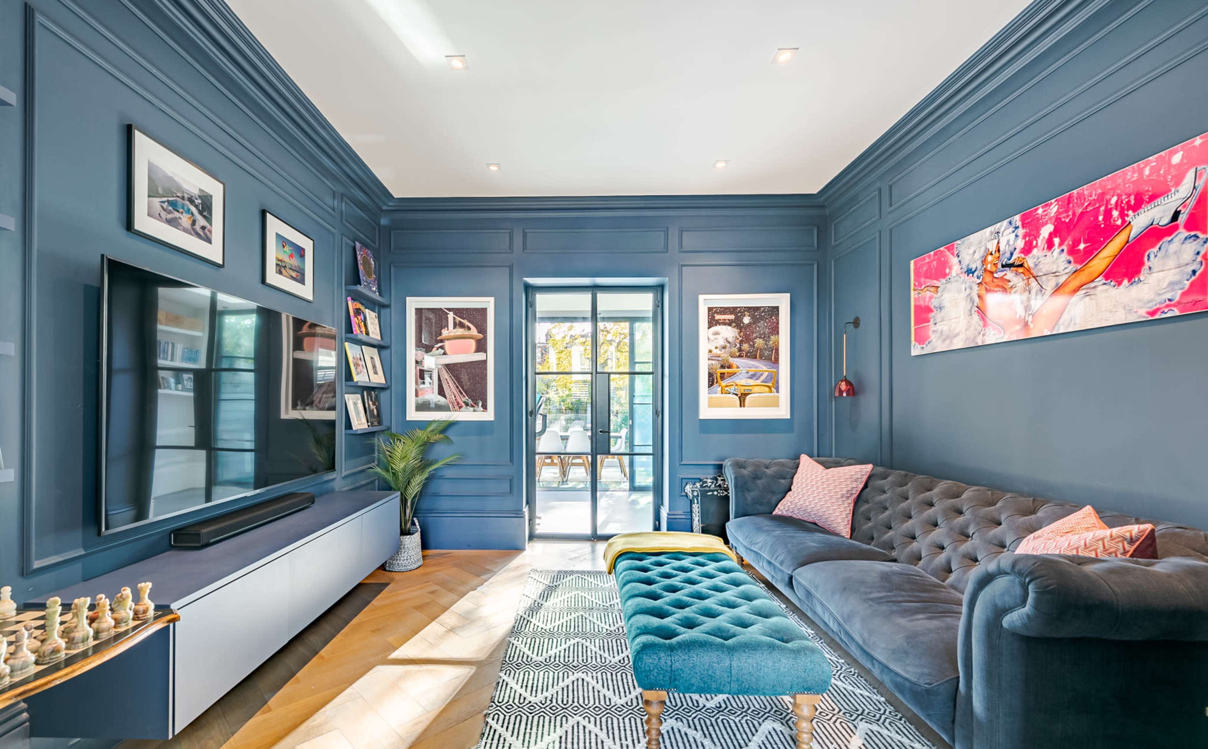 The image shows a modern living room with blue walls, a large flat-screen TV, a tufted sofa, and artwork hanging on the walls, leading to a door that opens to an outdoor space.