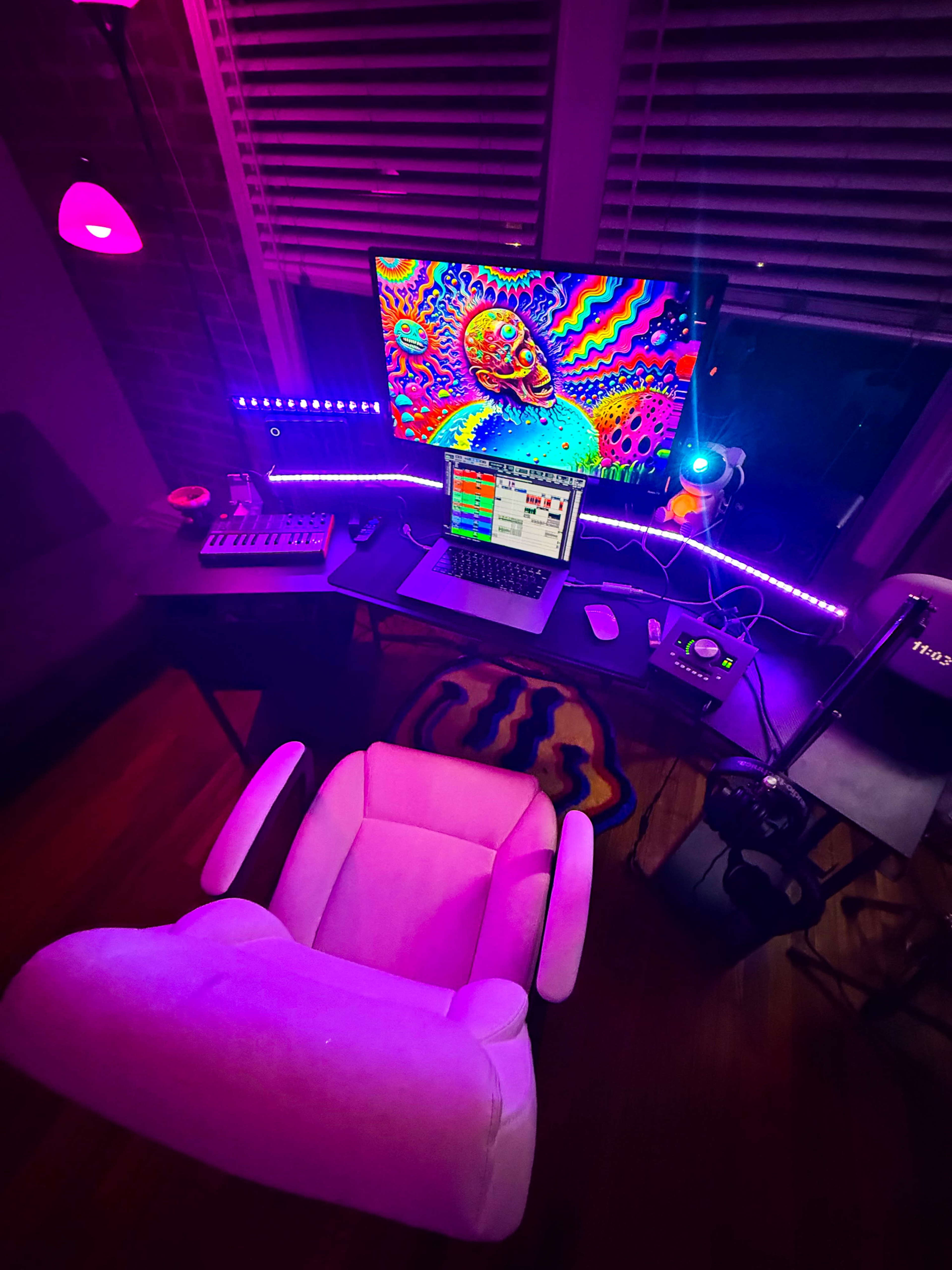 A colorful home office setup features a large monitor displaying vibrant digital art, a laptop, and ambient lighting in shades of purple.
