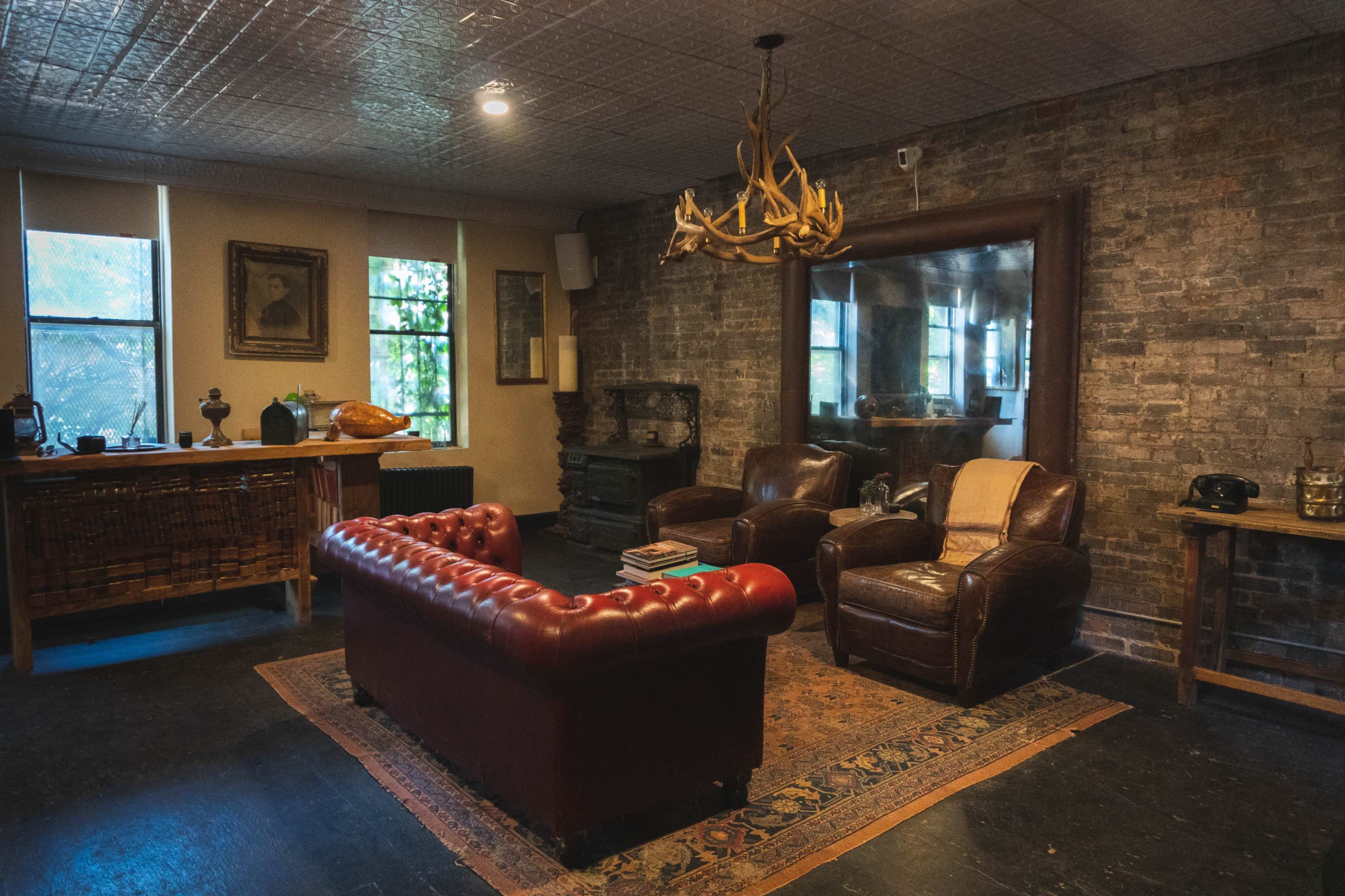 A cozy living room features a vintage leather sofa and armchairs, with a large mirror, a rustic chandelier, and exposed brick walls.