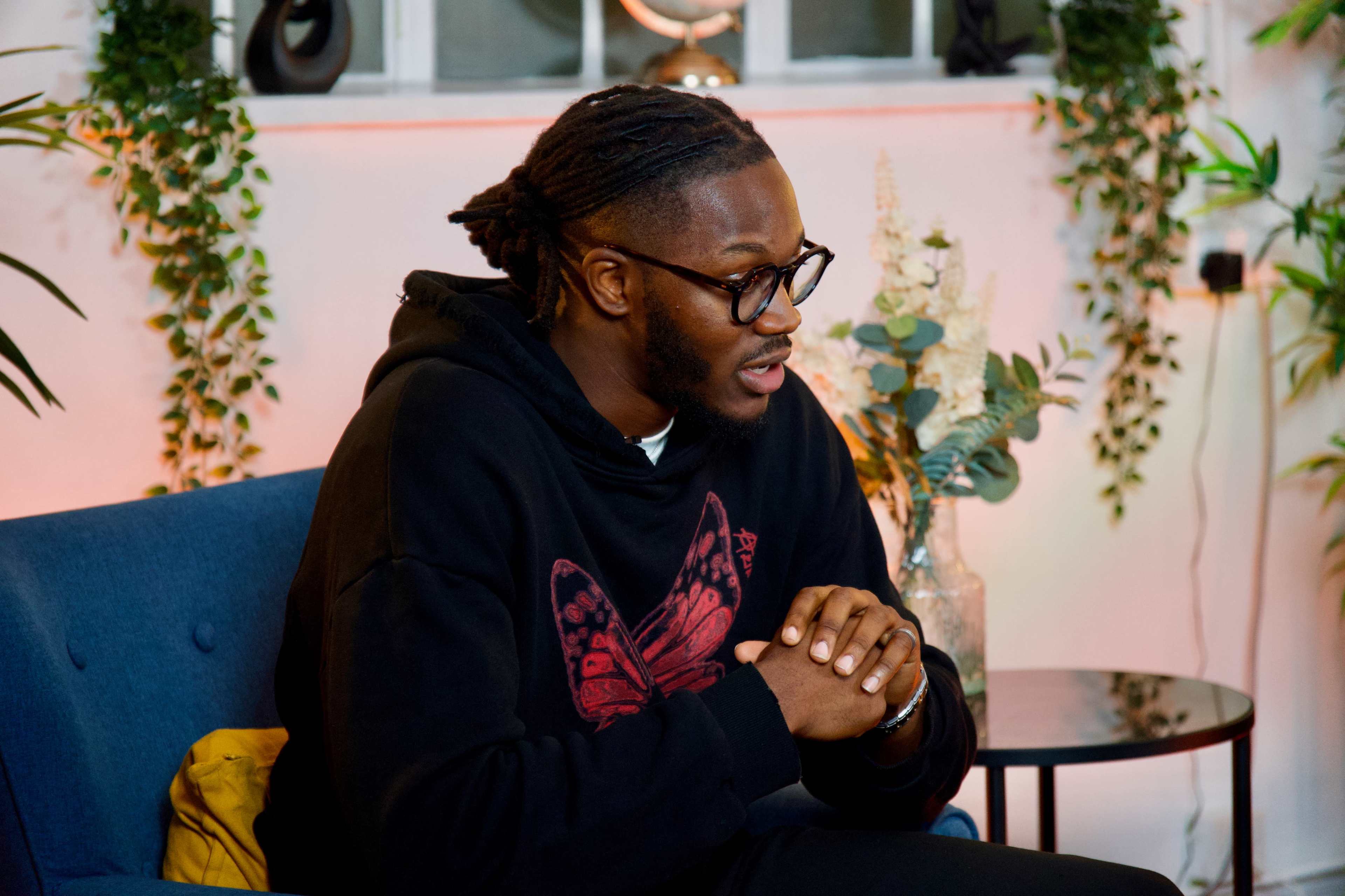 A man wearing glasses and a black hoodie sits on a blue sofa, engaging in conversation amidst plants and soft lighting.