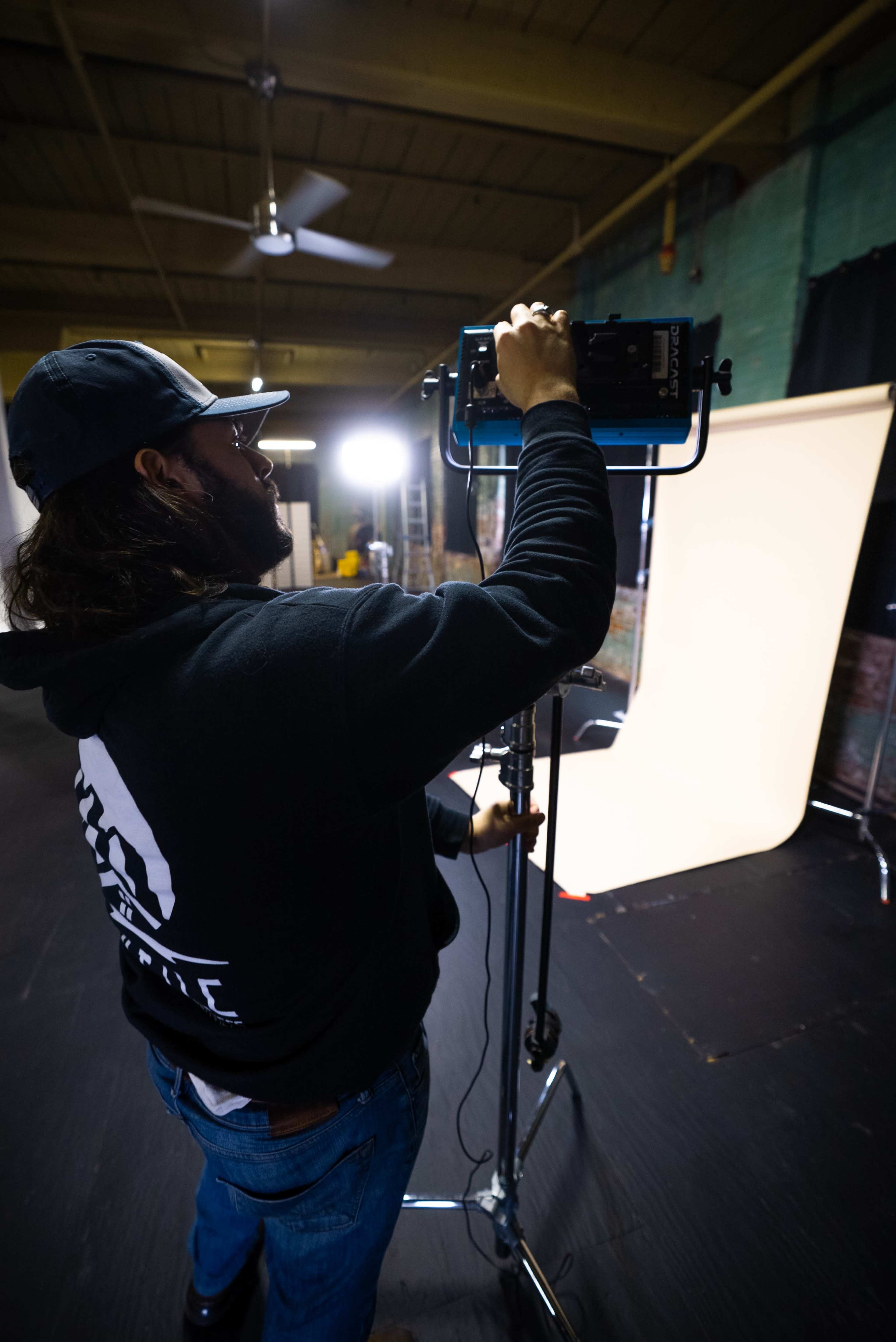 A person adjusts a photography light on a stand in a studio space with a backdrop set up.
