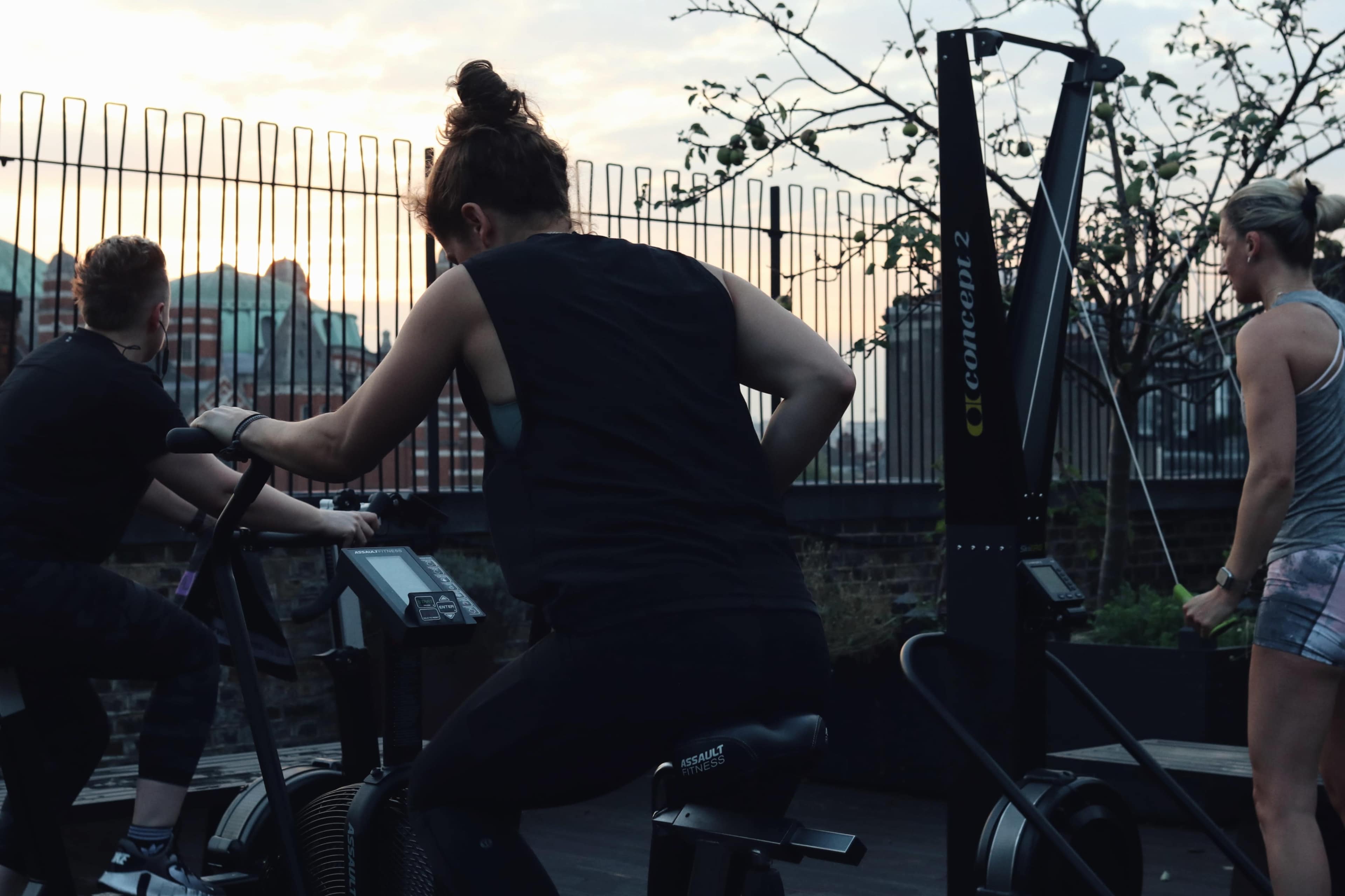 Three individuals are exercising on fitness equipment outdoors at sunset on a rooftop.