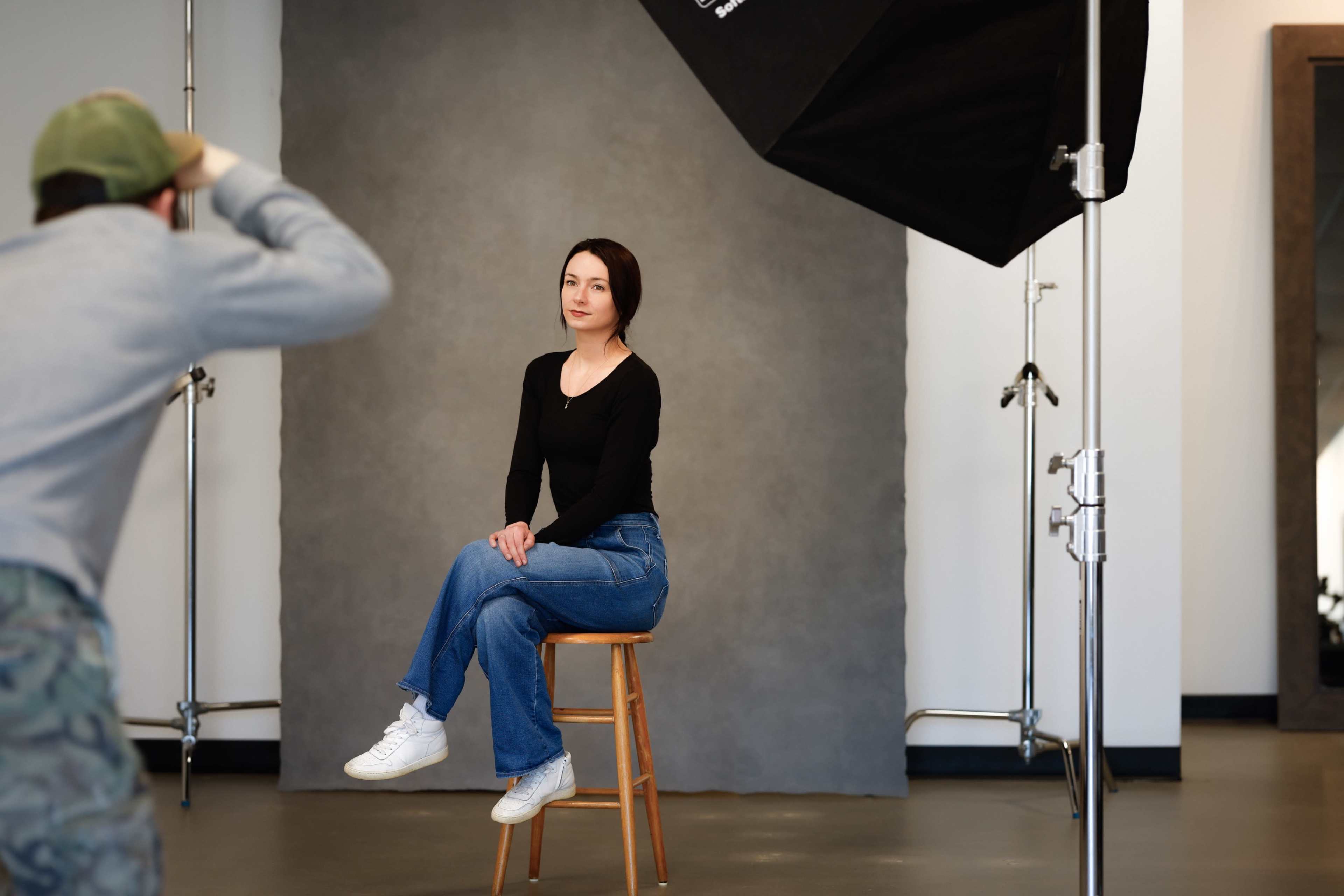 A woman sits on a stool in a photo studio as a photographer captures her portrait.
