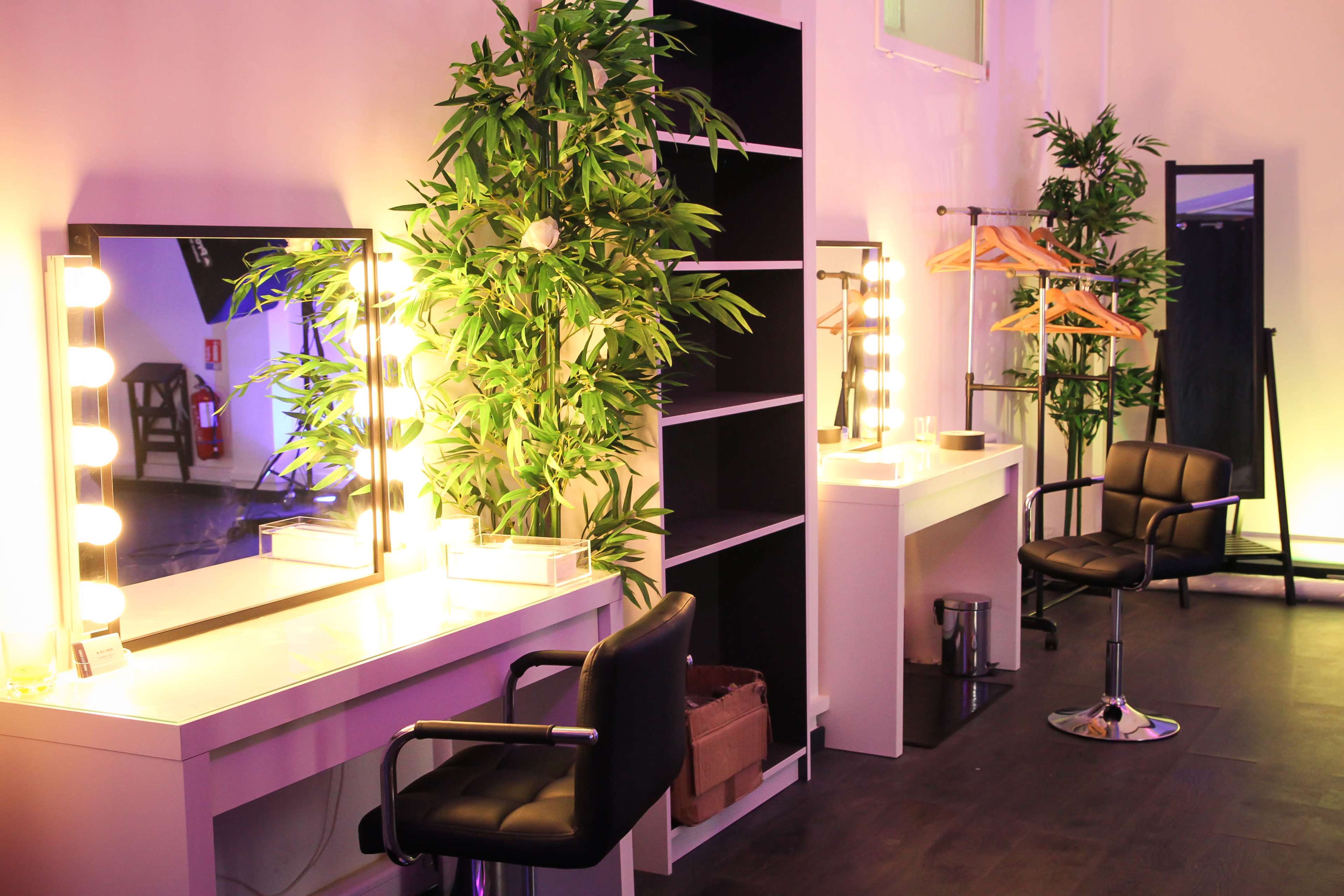 The image shows a brightly lit dressing room area featuring mirrors with lights, a grooming chair, a clothing rack, and decorative plants.