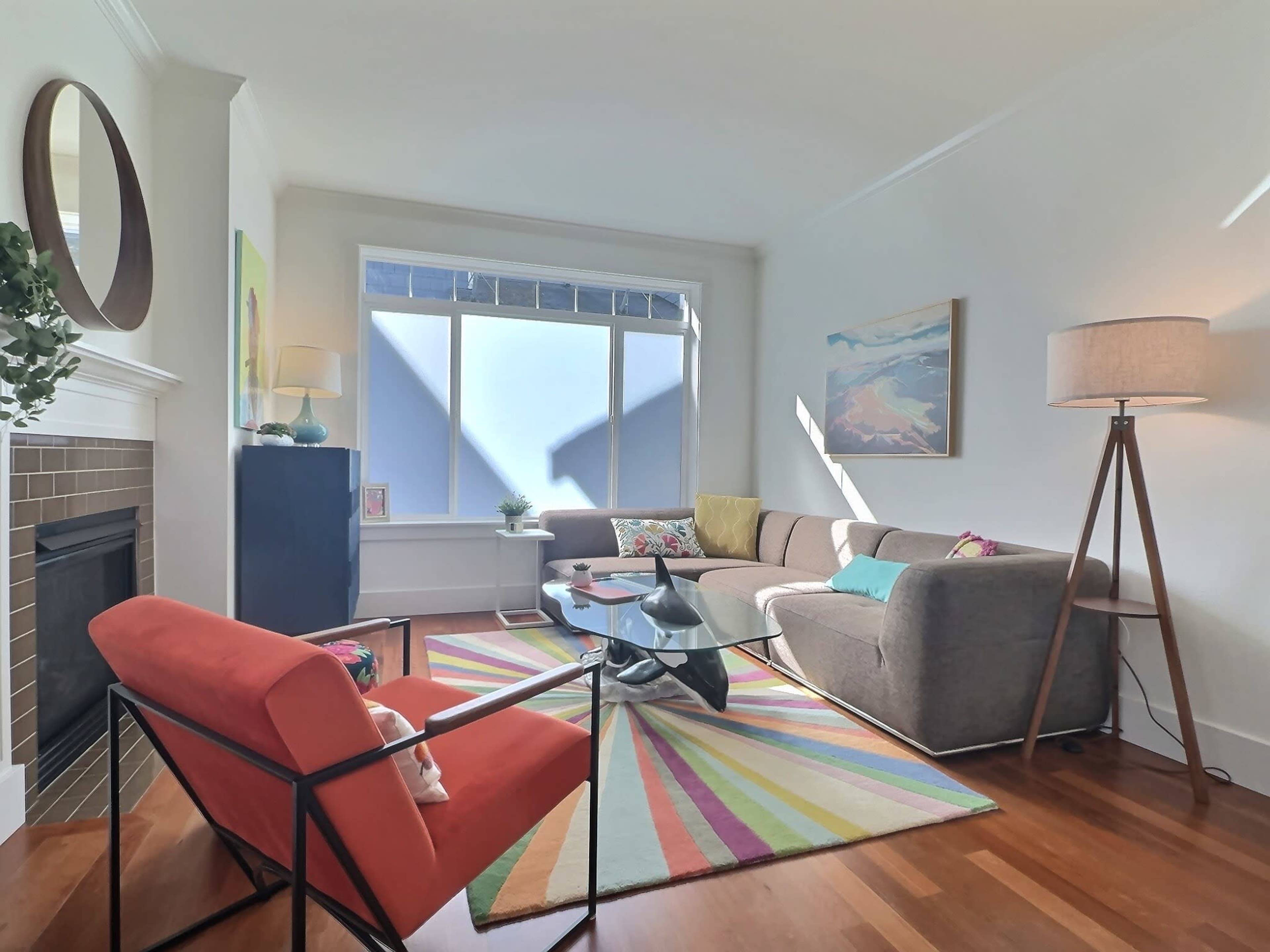 The image shows a brightly lit living room featuring a colorful area rug, a gray sofa, an orange chair, a coffee table, and large windows with natural light.