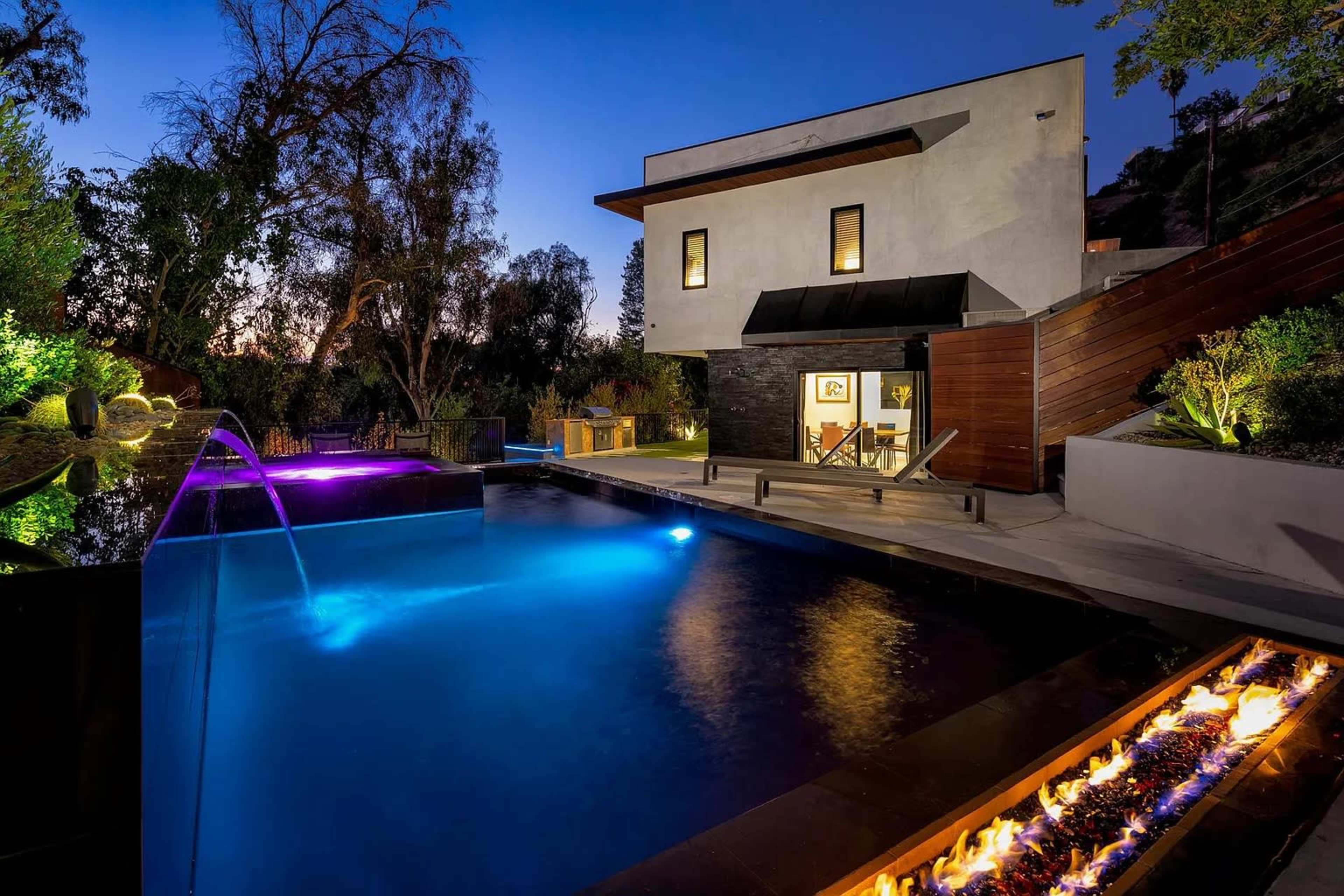 A modern house is set beside a swimming pool with colorful LED lighting at dusk, surrounded by landscaped greenery.