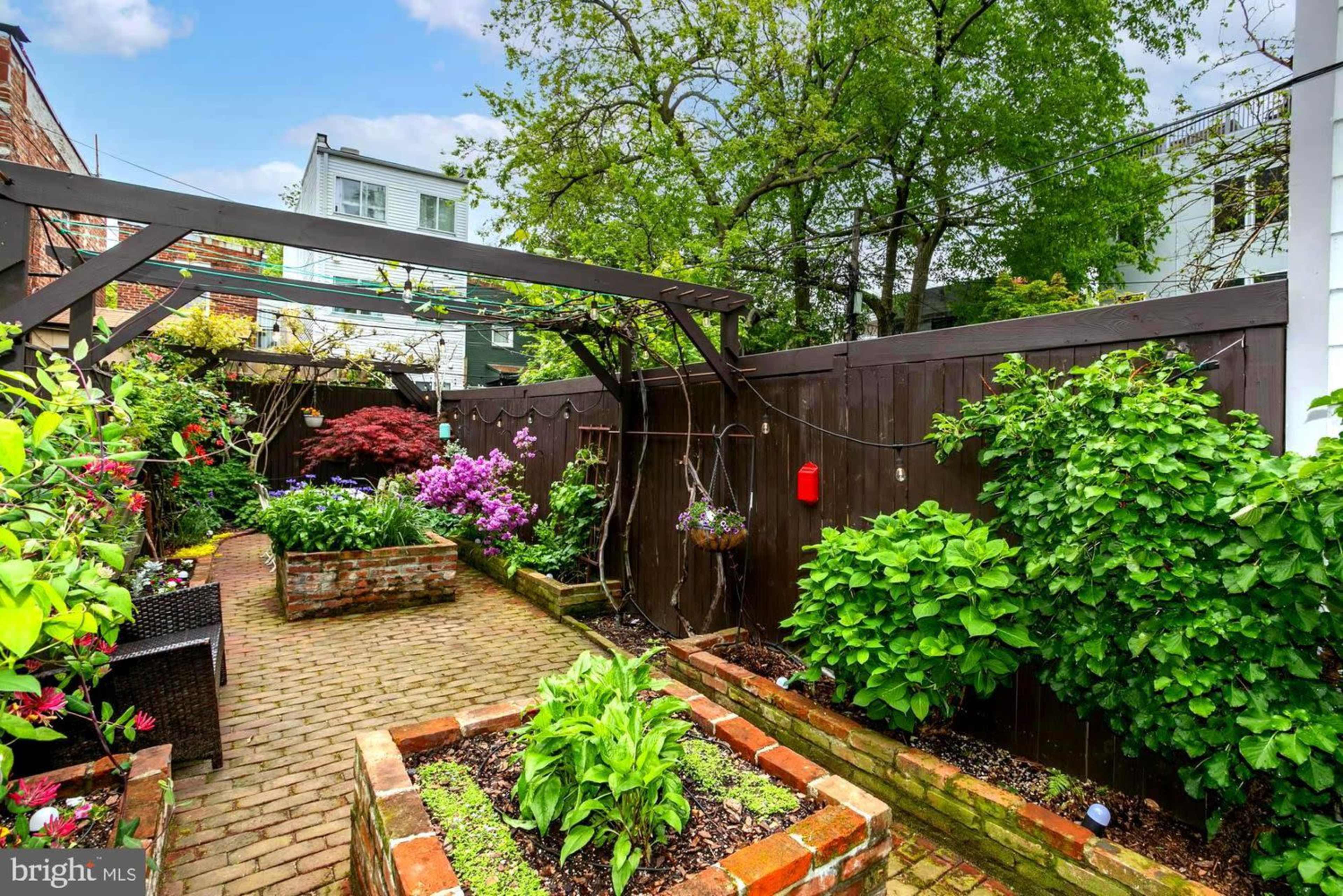 A well-maintained backyard garden features brick pathways, raised flower beds with various plants, and a pergola draped with greenery.