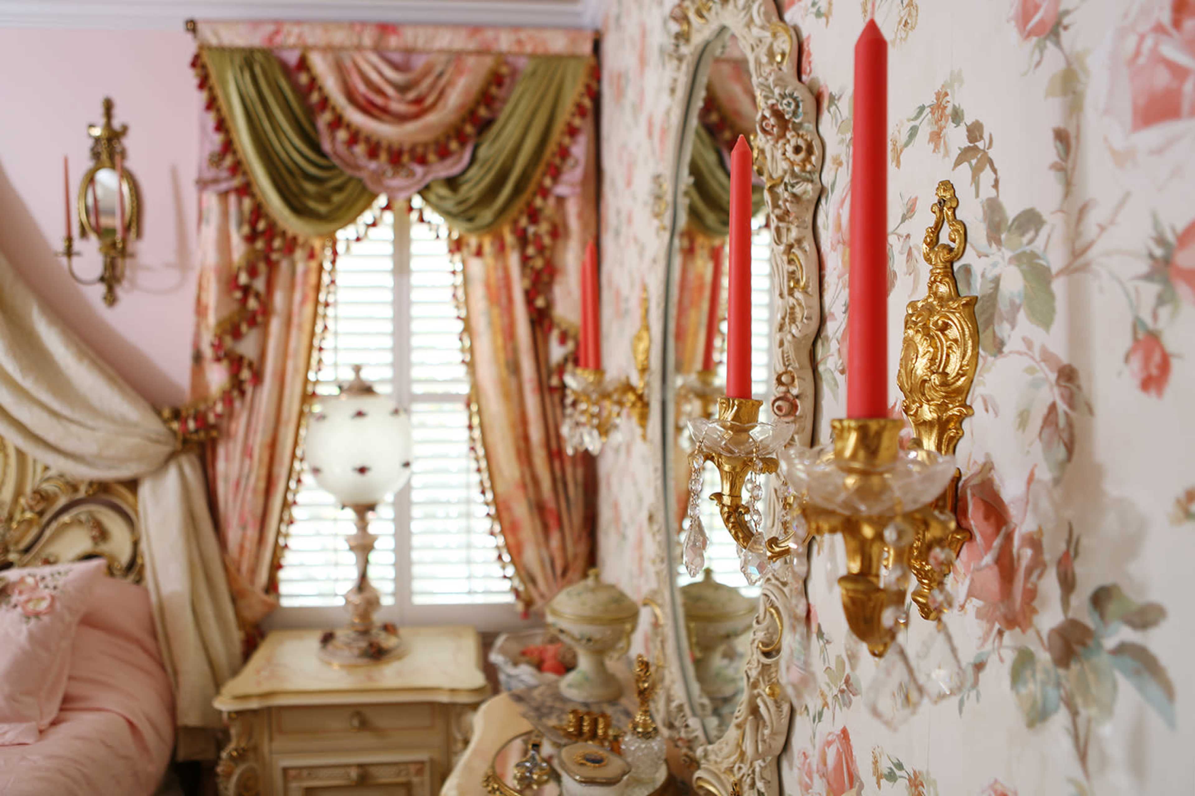 A elegantly decorated bedroom features floral wallpaper, ornate mirrors, and candle sconces flanking a window covered with draped curtains.