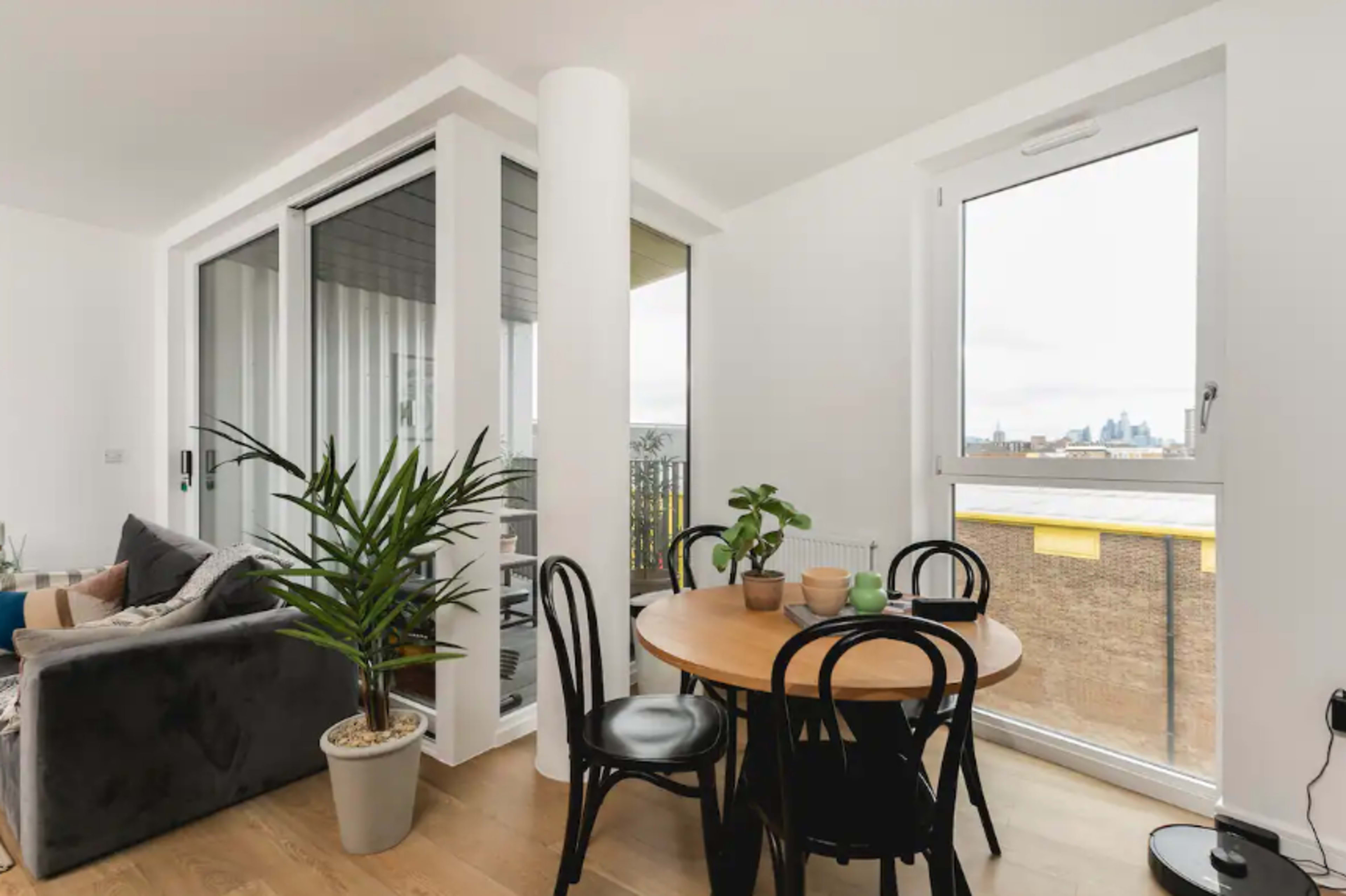 A living area features a round dining table surrounded by black chairs, with a plant in a pot and large windows showcasing an outdoor view.