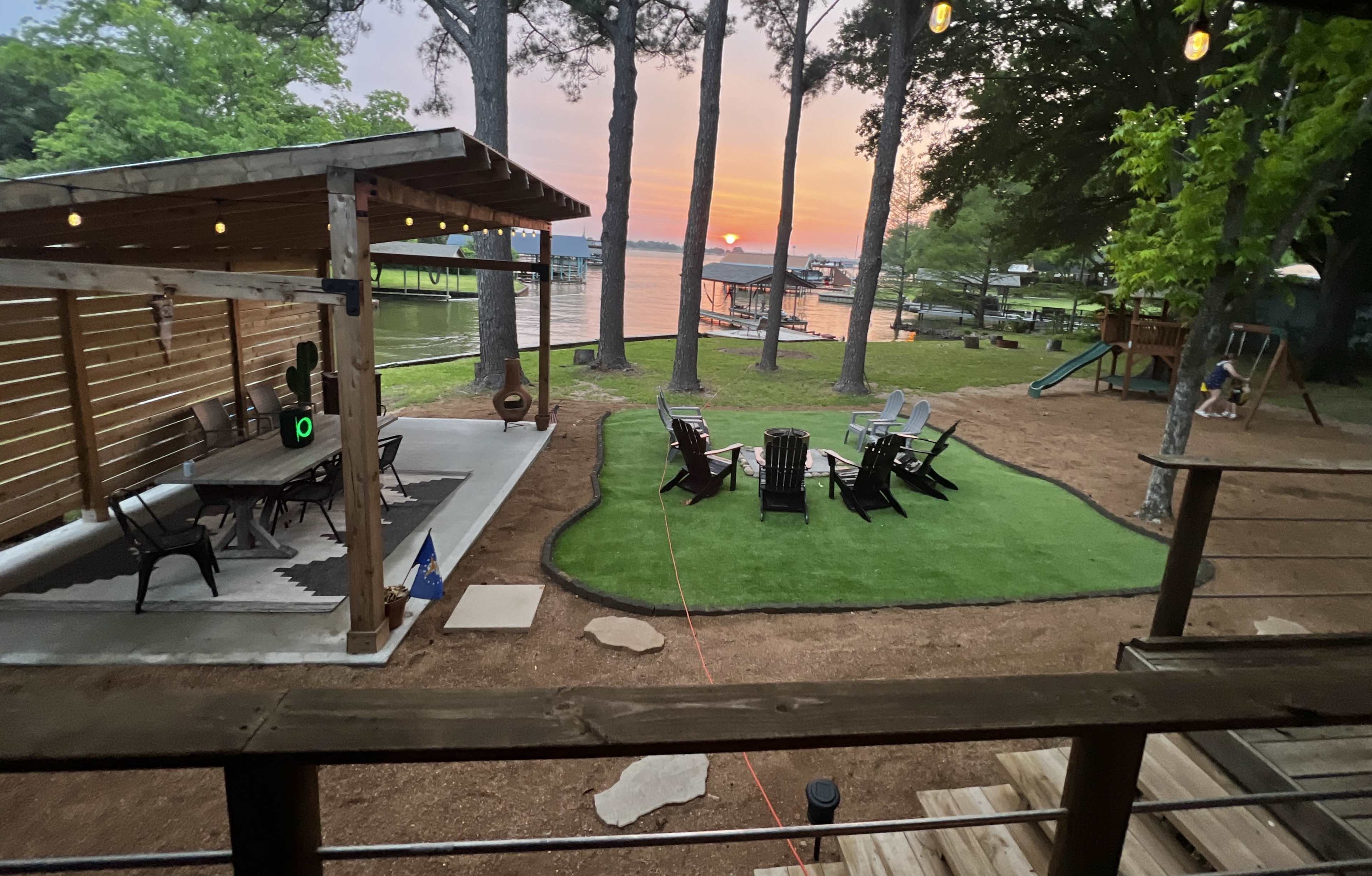 A lakeside setting with a covered patio, multiple lawn chairs arranged on a grassy area, and a view of the sunset reflecting on the water.