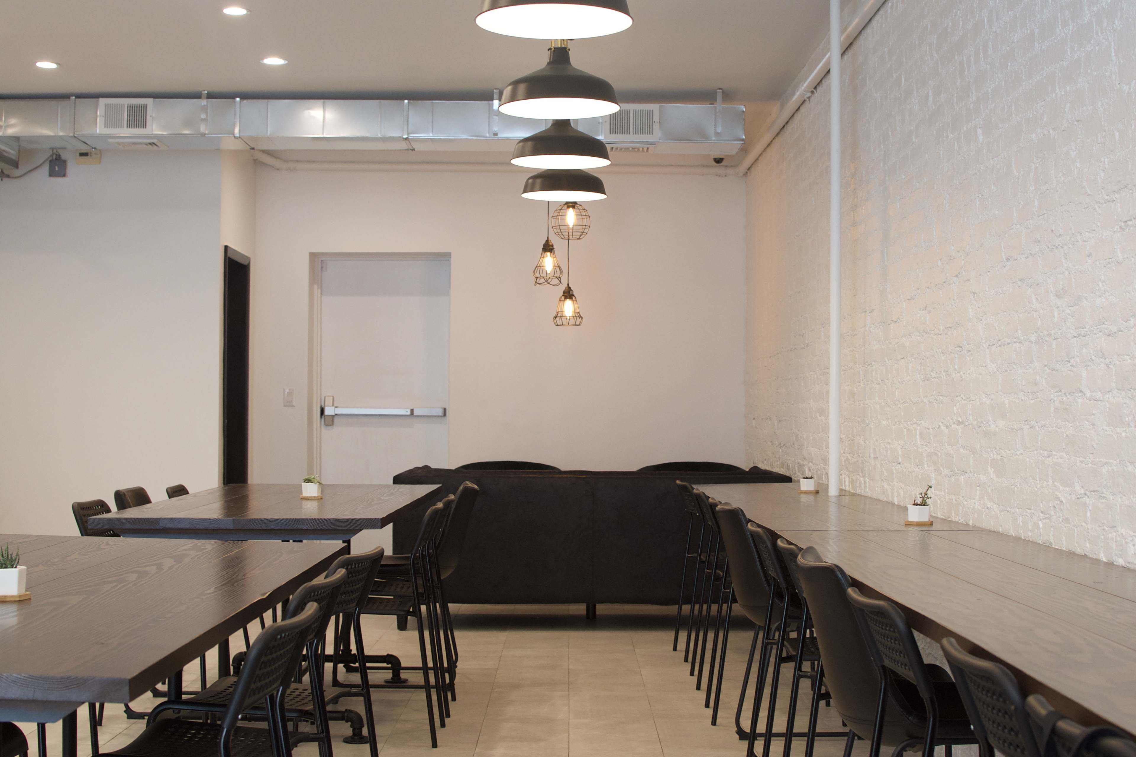 A modern dining area with several long wooden tables, black chairs, and pendant lighting hanging from the ceiling, along with a couch against a white brick wall.
