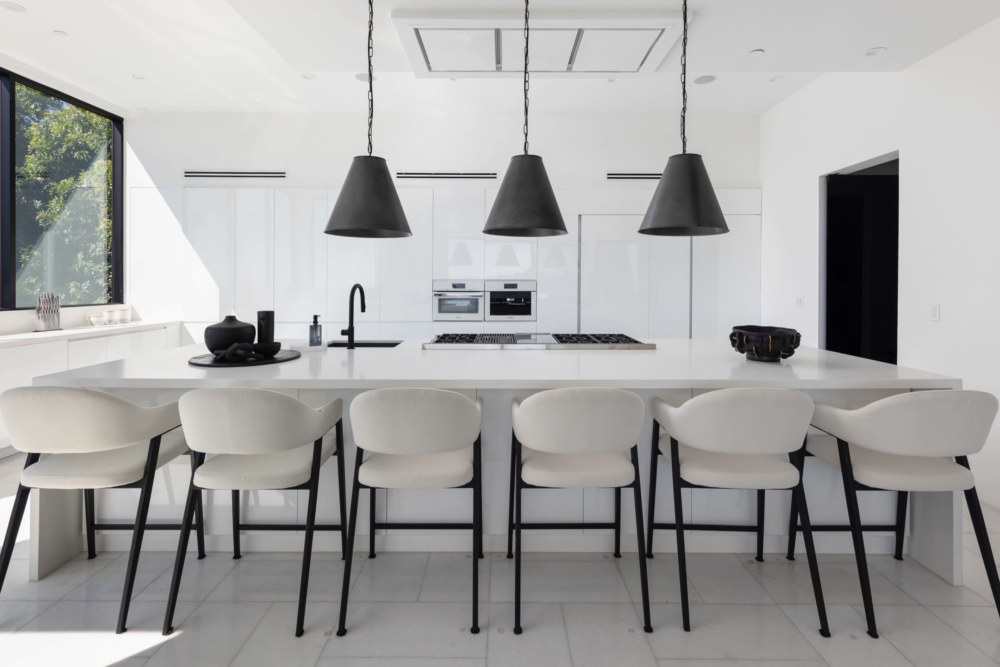 A modern kitchen features a long white island with six upholstered stools, minimalist cabinetry, and three large pendant lights hanging above.