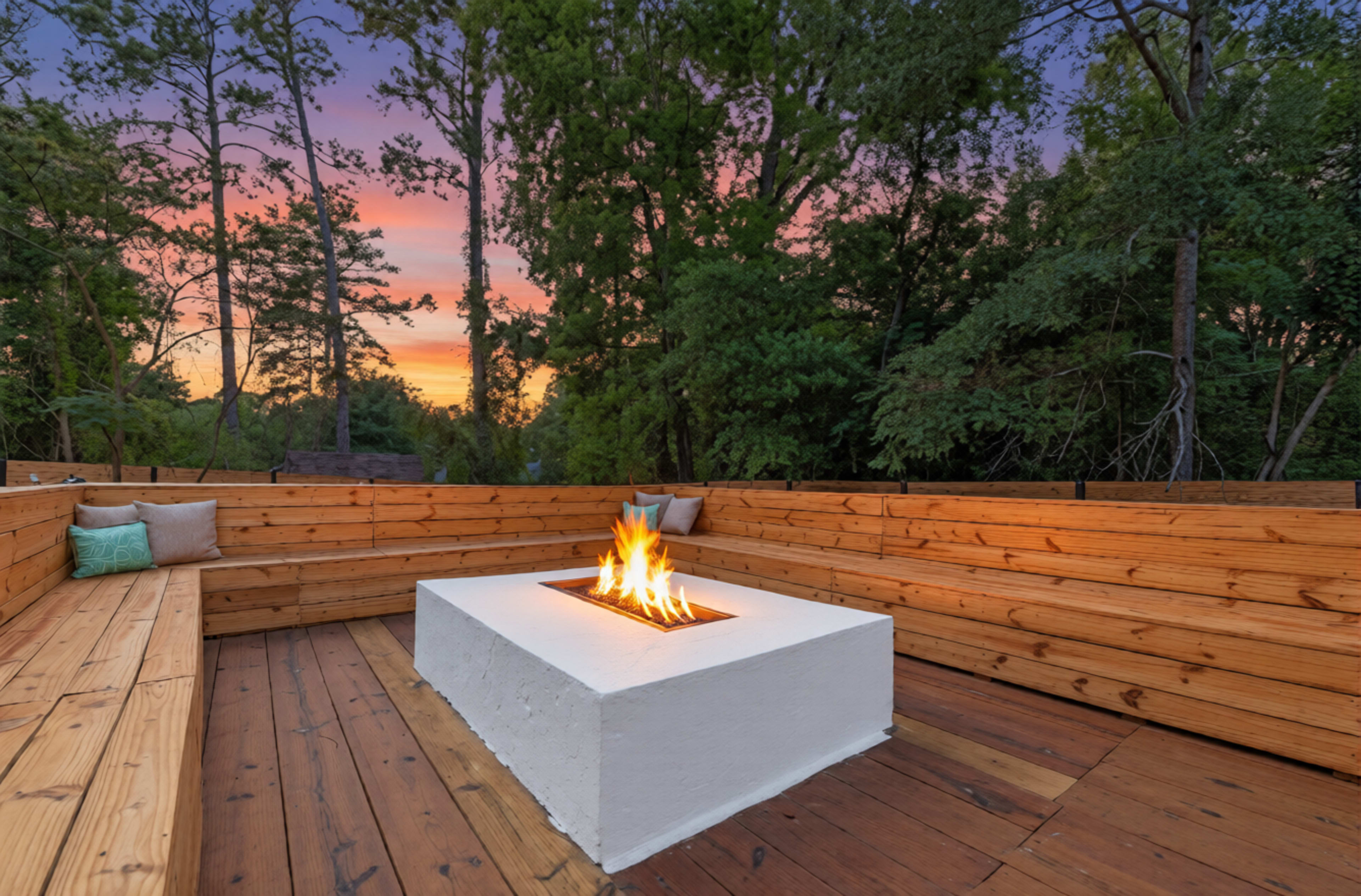 A rectangular fire pit with flames is situated in the center of a wooden deck, surrounded by wooden benches, under a colorful sunset sky and tall trees.