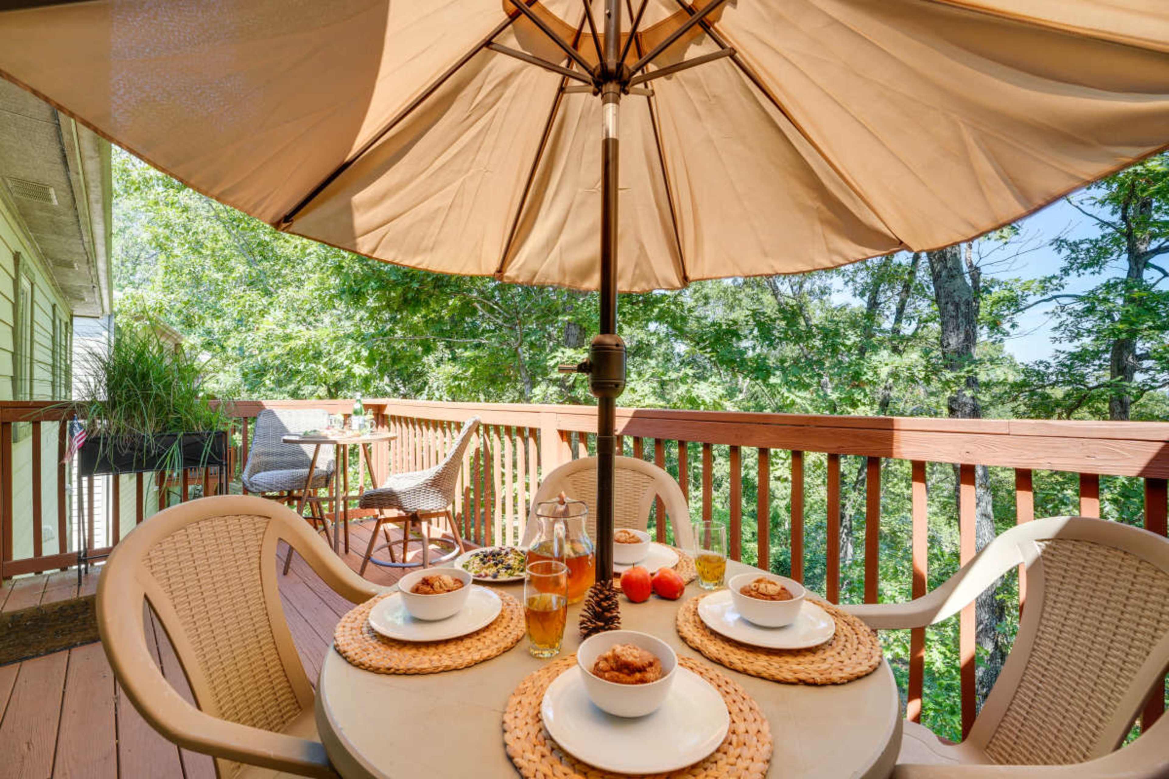 A table set with bowls of food and drinks is placed under a large umbrella on a deck overlooking greenery.