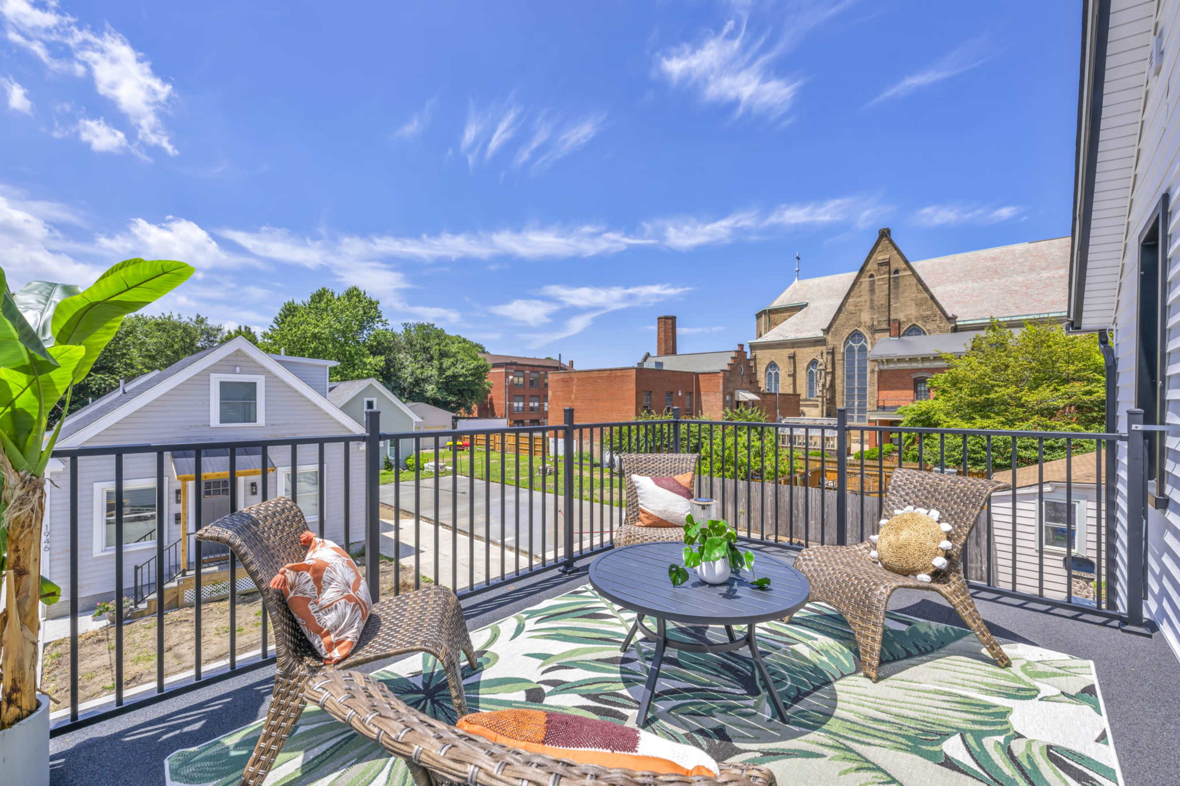A balcony features two woven chairs and a table with potted plants, overlooking a neighborhood with a church and buildings in the background.