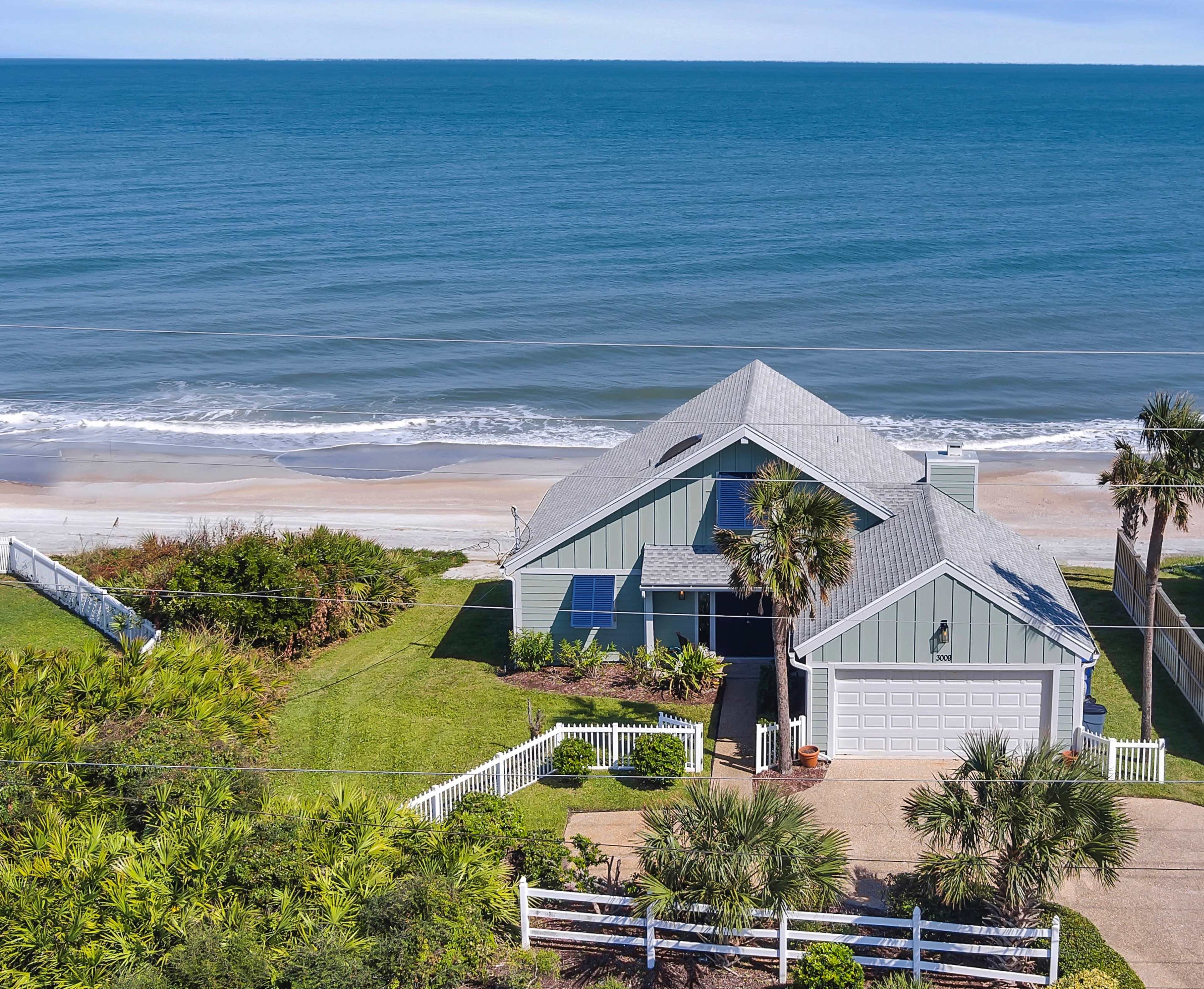 A coastal house with a gray roof and a beach visible in the background, surrounded by greenery and a white picket fence.
