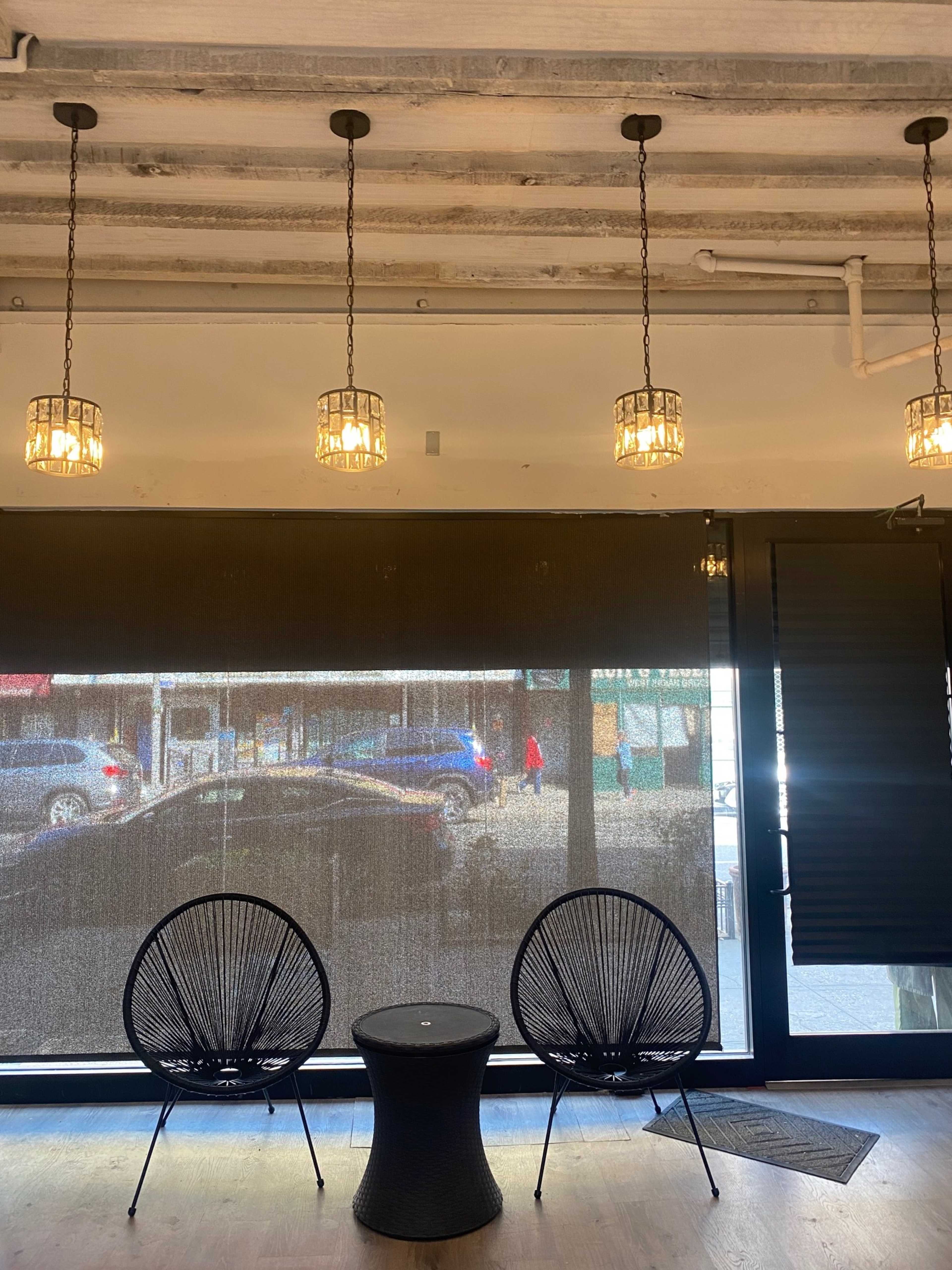 A modern interior with two black chairs facing a small table, illuminated by hanging light fixtures above and a large window displaying an outdoor street scene.