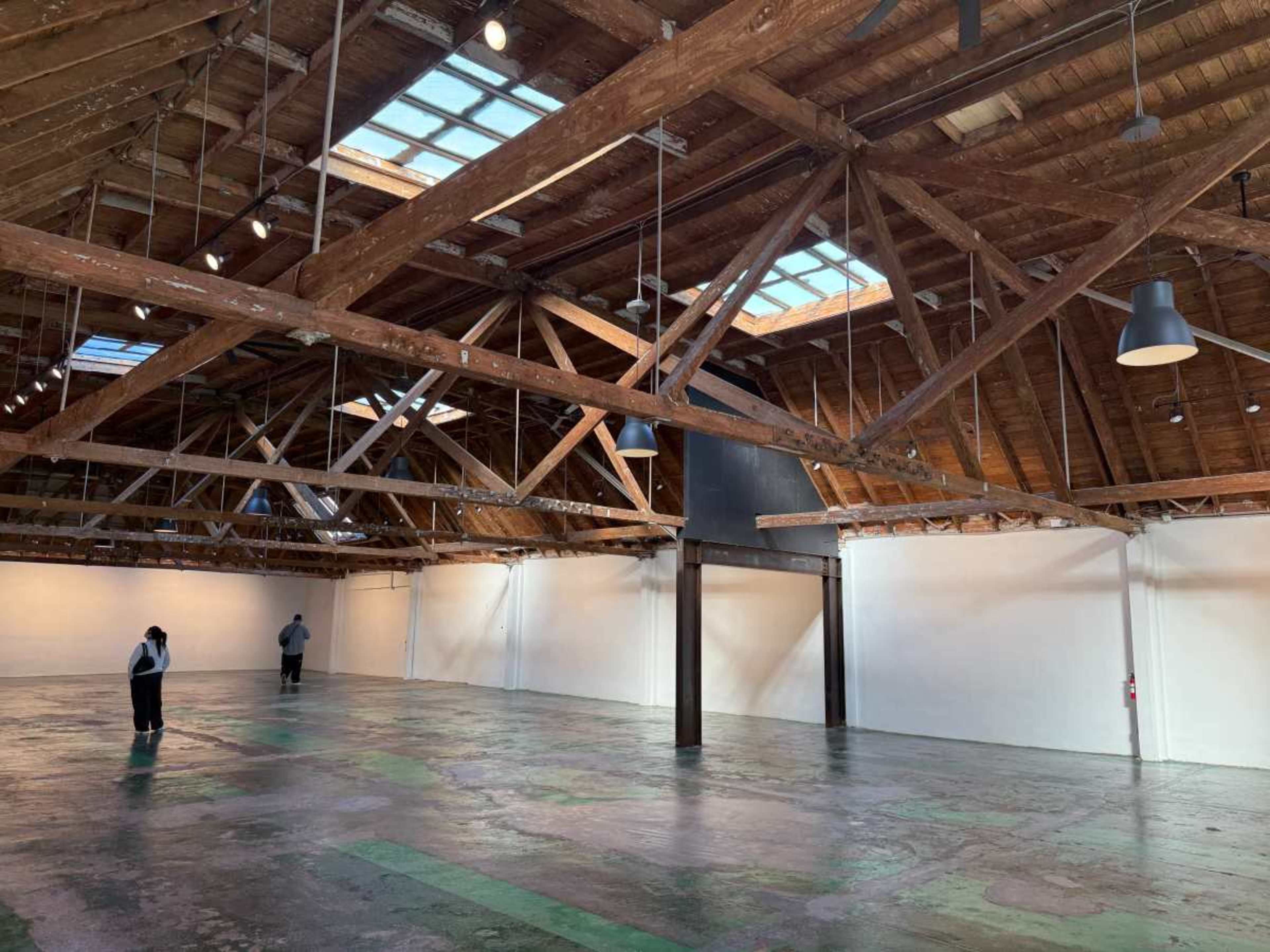 The image shows an empty industrial space with exposed wooden beams, large skylights, and a polished concrete floor.