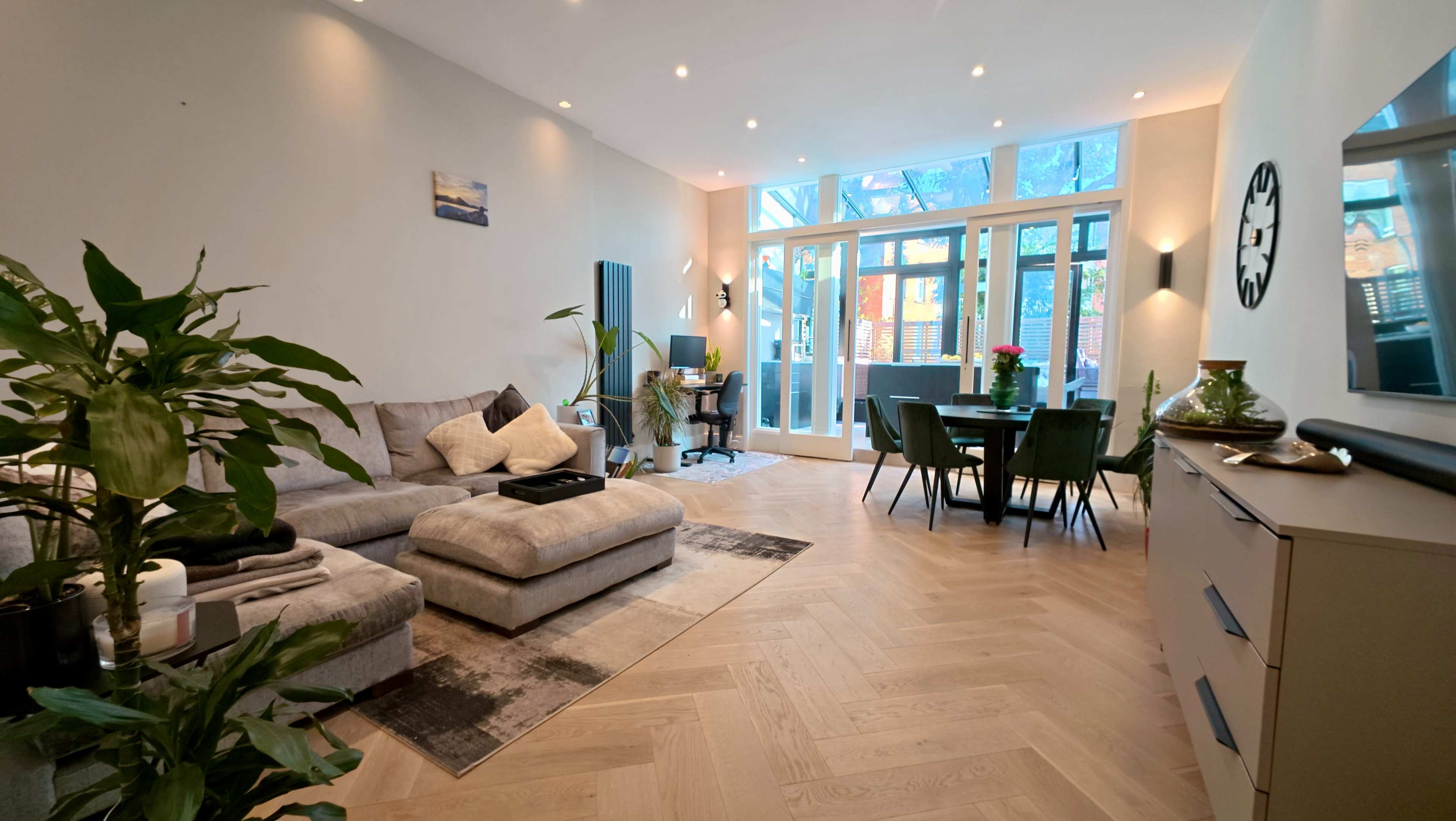 The image shows a modern living room featuring a gray sectional sofa, a dining area with green chairs, and large windows offering natural light.