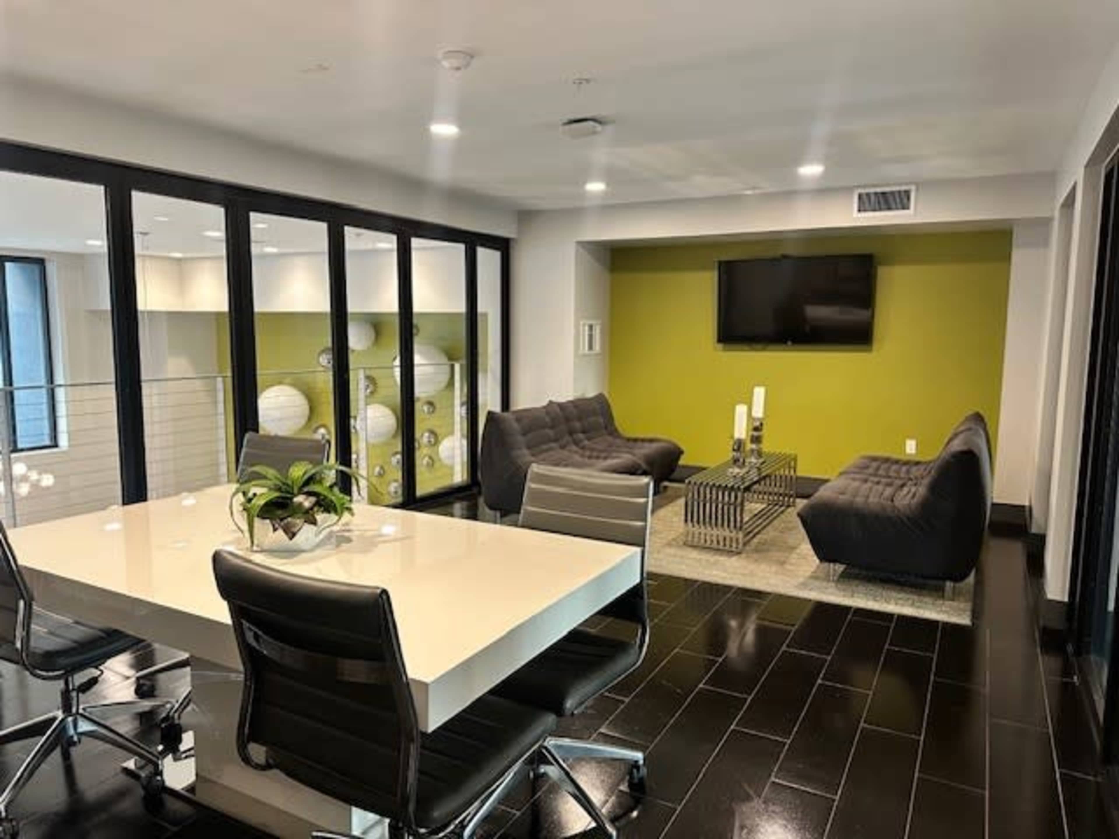 The image shows a modern office lounge with sleek furniture, including two gray sofas and a glass table, set against a green accent wall and a television.