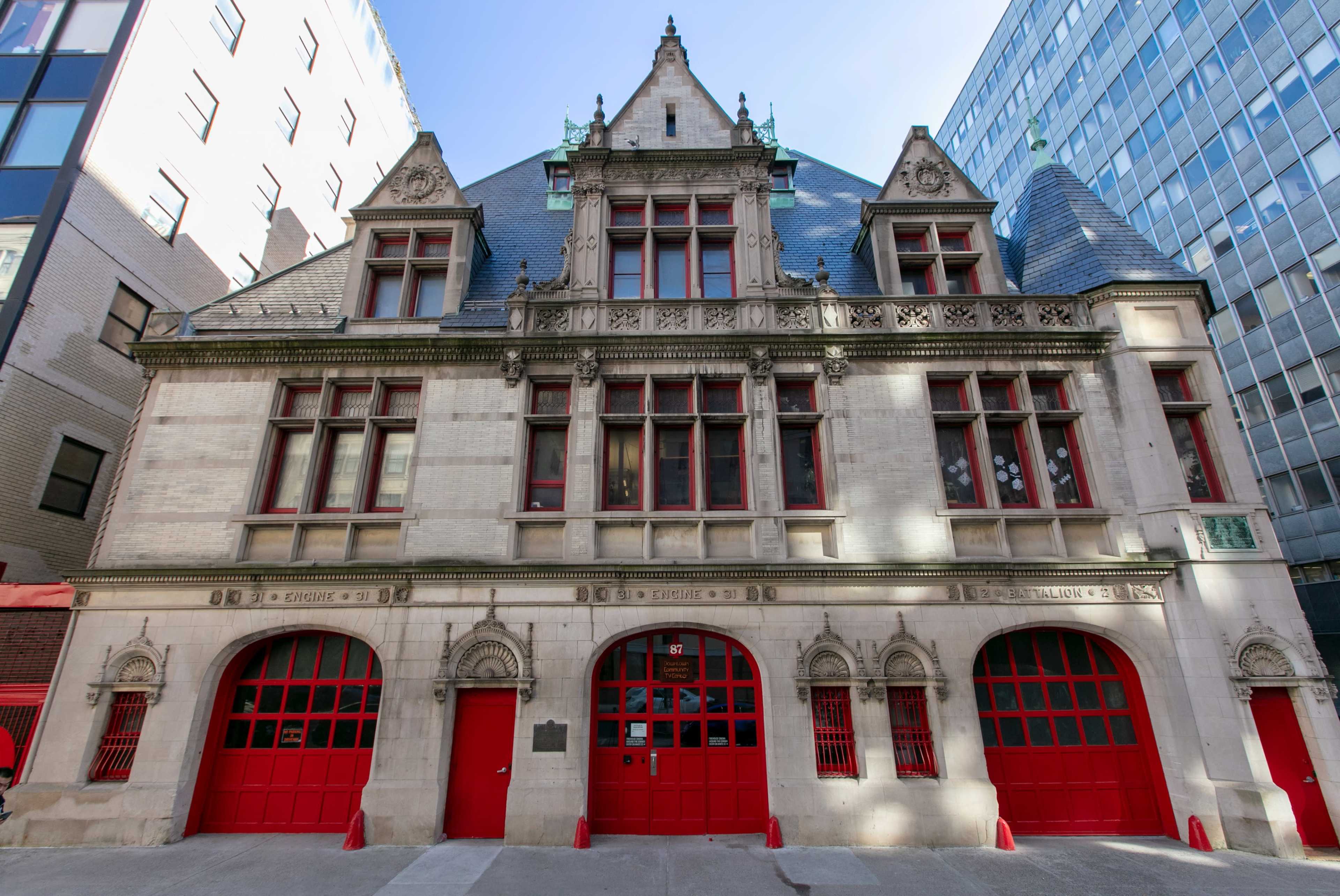 The image shows a historic building with a peaked roof, intricate architectural details, and bright red doors, situated between two modern structures.