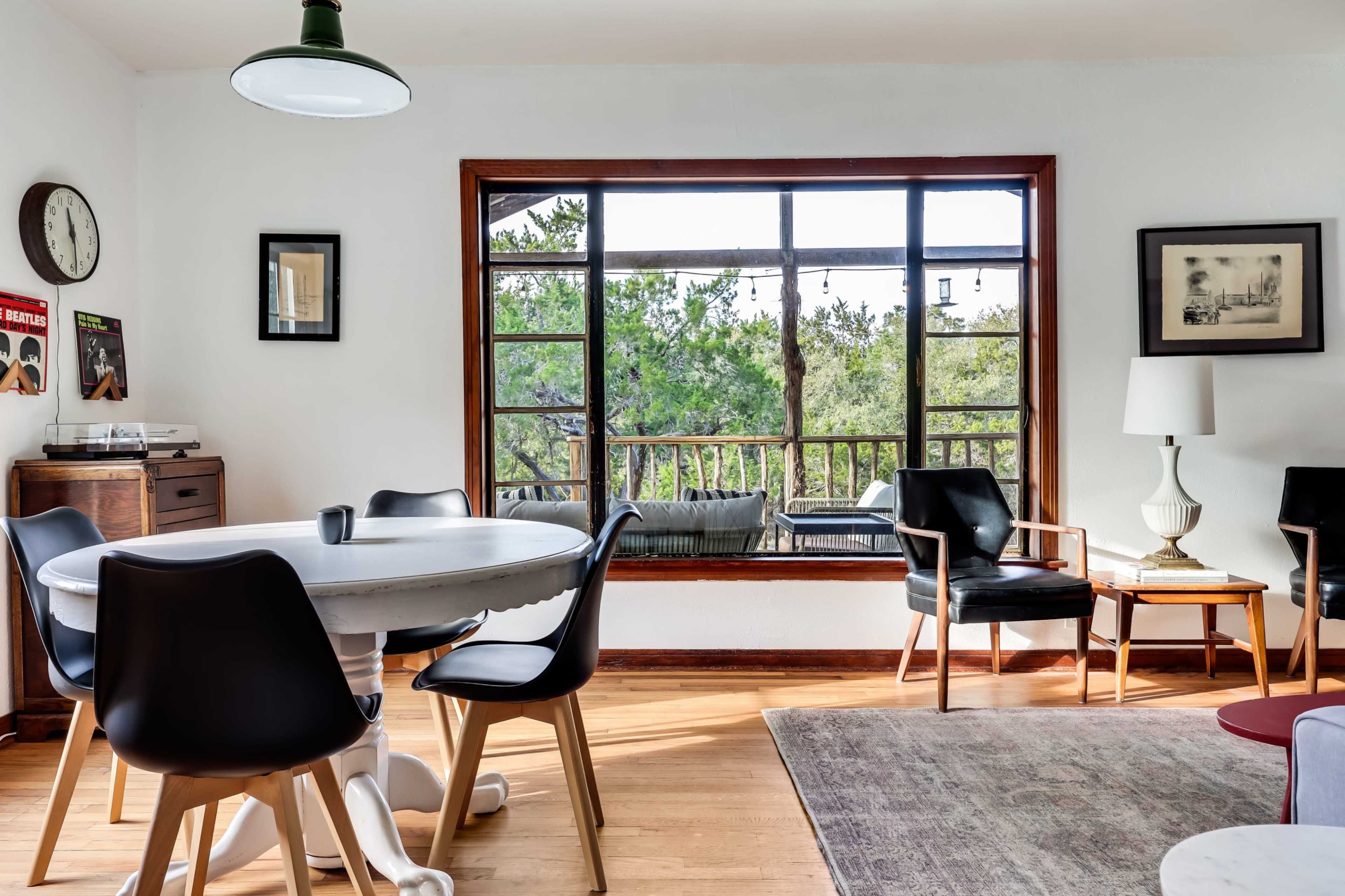 A dining area features a round white table surrounded by black chairs, with a large window overlooking greenery and a small outdoor space.