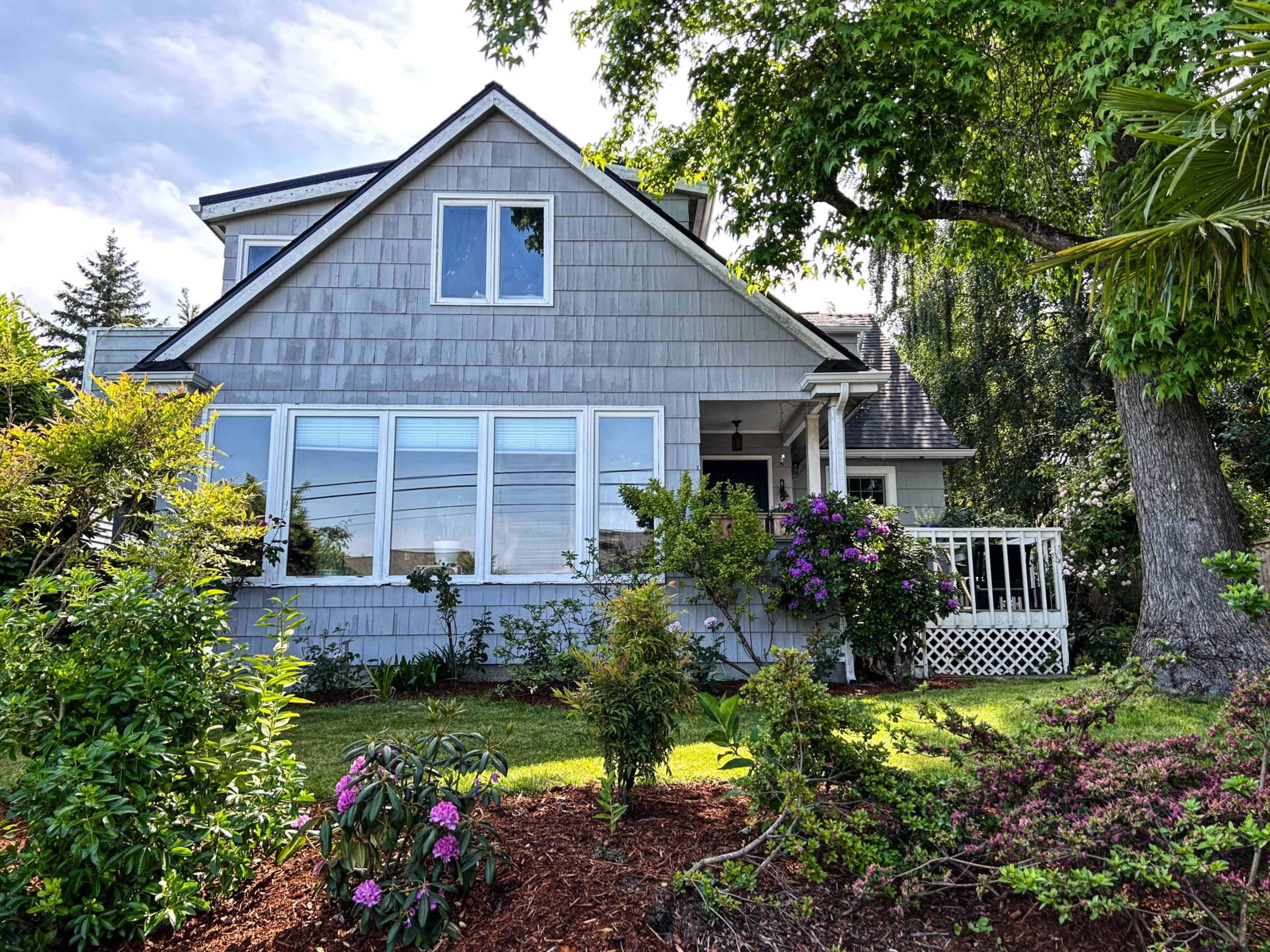 A two-story house with gray shingles features large windows, a front porch, and is surrounded by a landscaped yard with various plants and flowers.