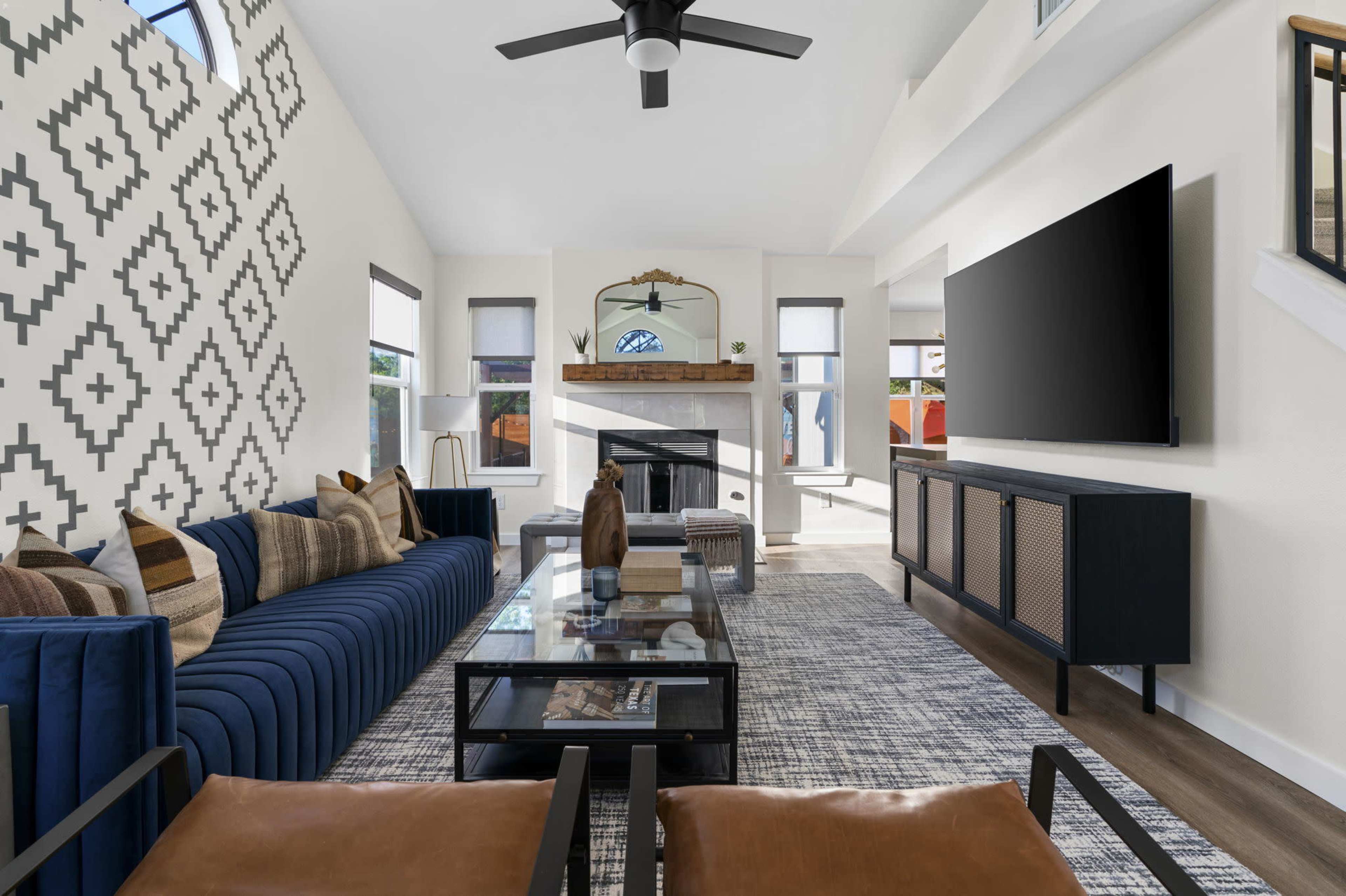A modern living room features a blue sofa, a glass coffee table, two brown leather chairs, and a fireplace with a mounted television above it.