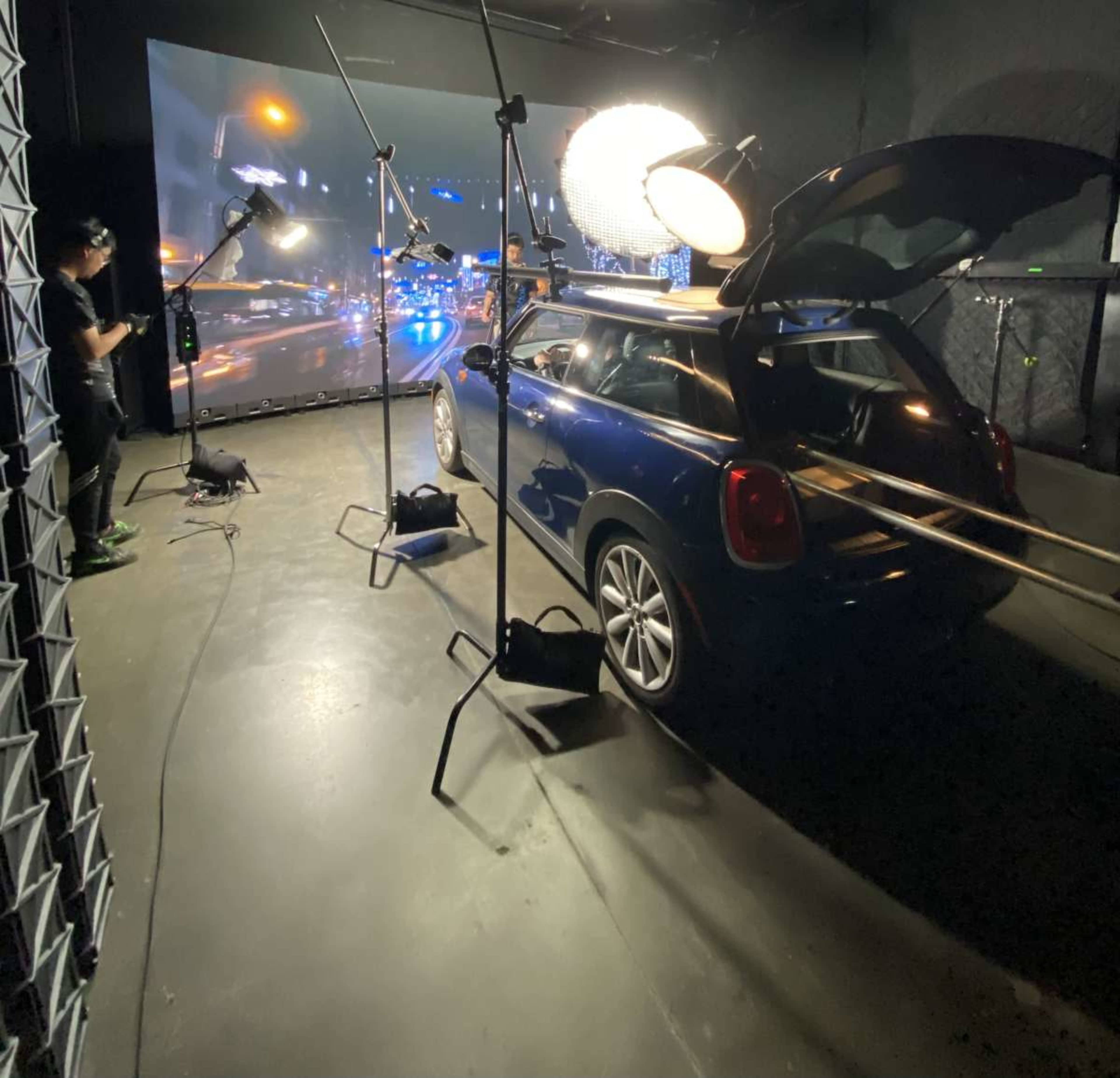 A blue mini car is parked in a studio with lighting equipment and a large screen displaying a cityscape in the background.