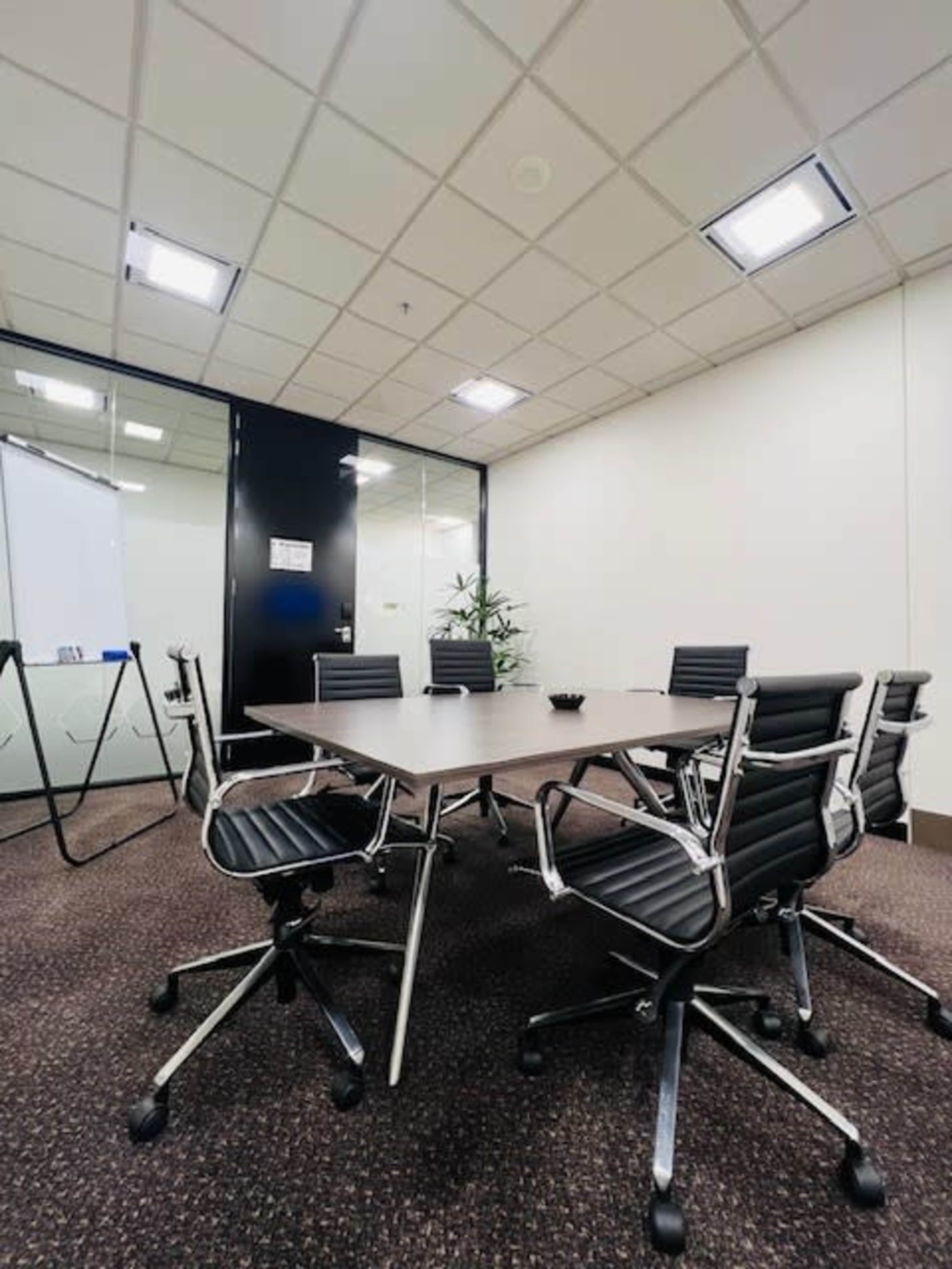 A modern conference room features a rectangular table surrounded by ergonomic chairs, with a whiteboard and a plant in the background.