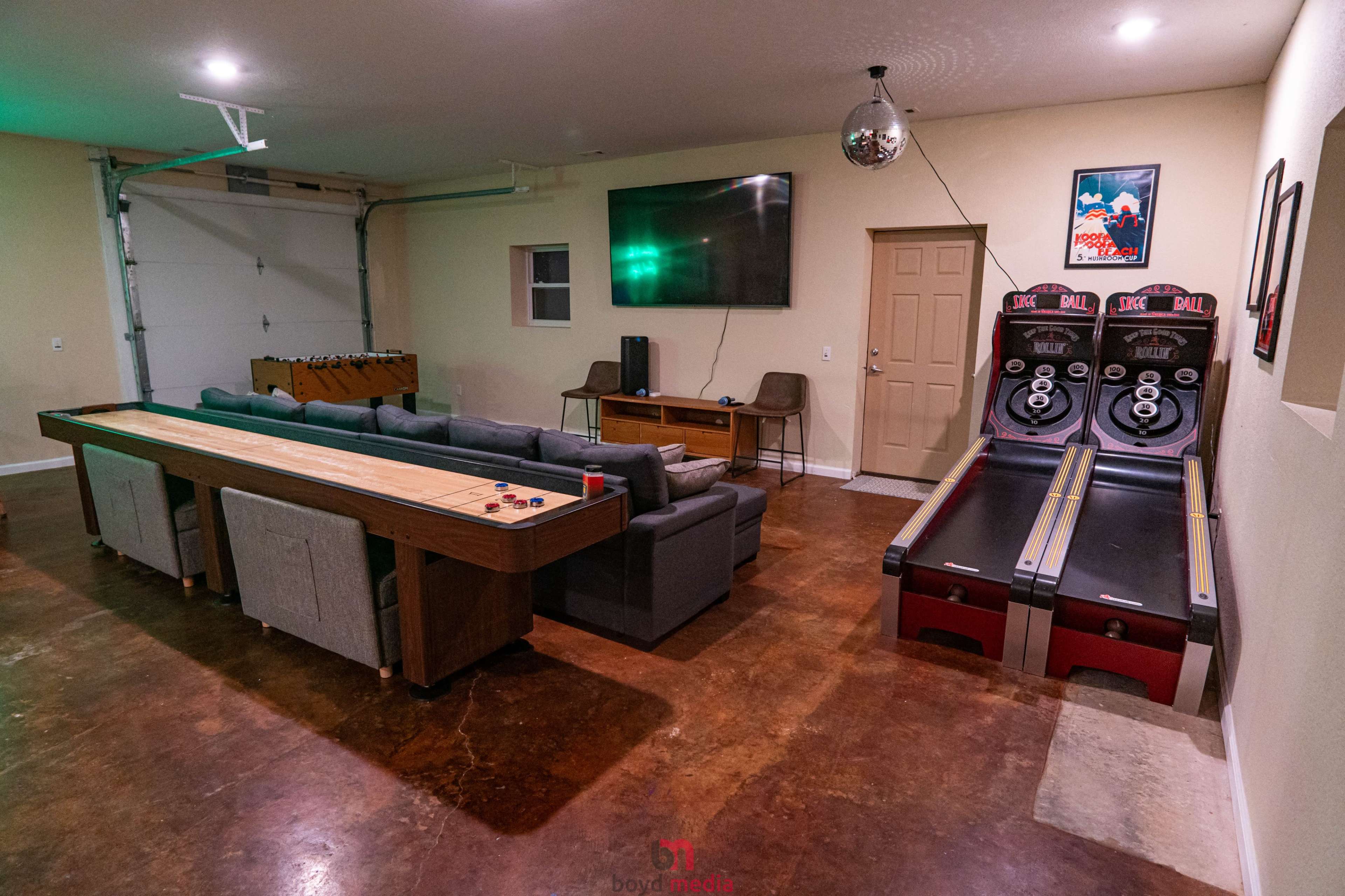 A recreational room with a couch, a television mounted on the wall, and two arcade-style skee-ball machines alongside a long wooden table.