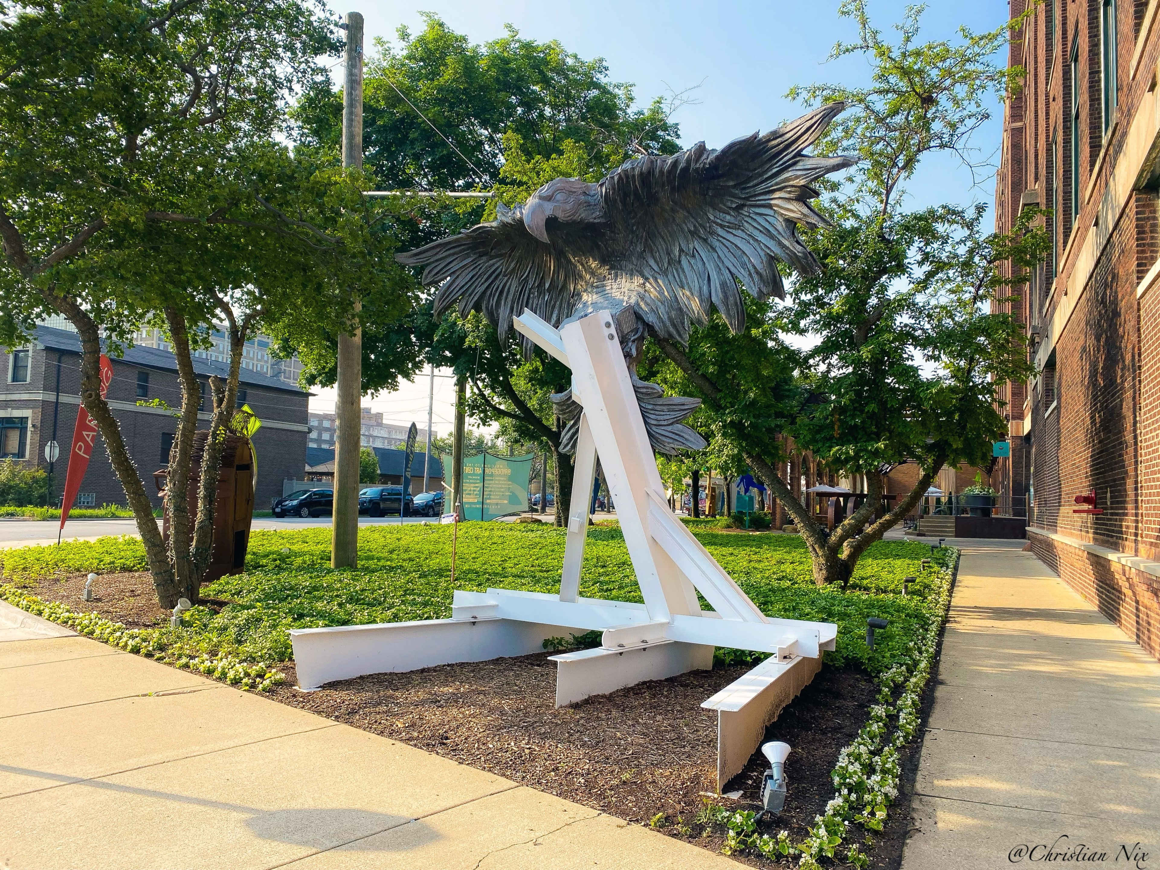 The image shows a large, metallic sculpture of an eagle taking flight, situated on a landscaped patch of ground surrounded by trees near a building.