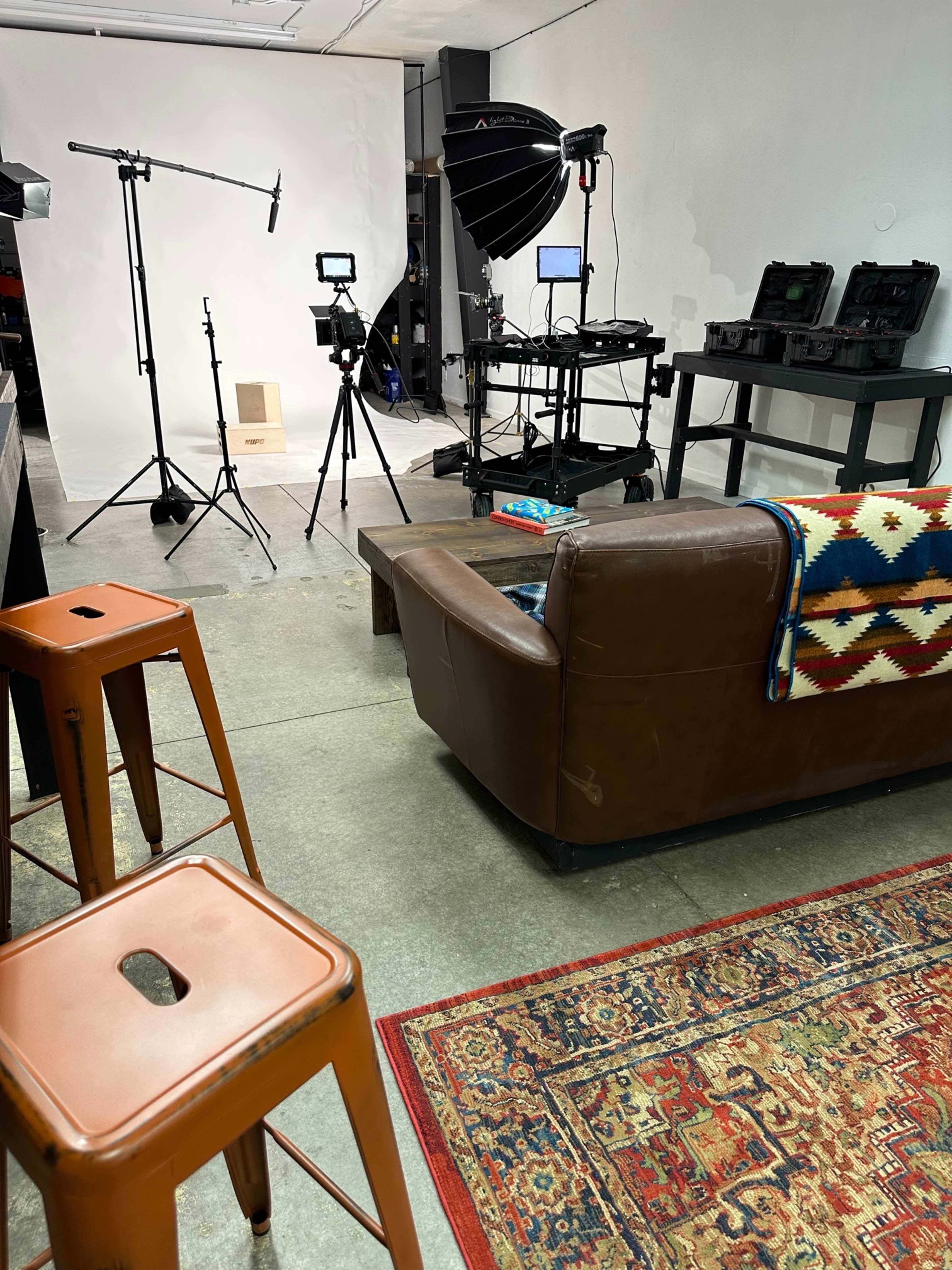 A well-lit indoor studio setup features a brown couch, metal bar stools, and various video equipment, including lights and cameras, arranged against a plain backdrop.