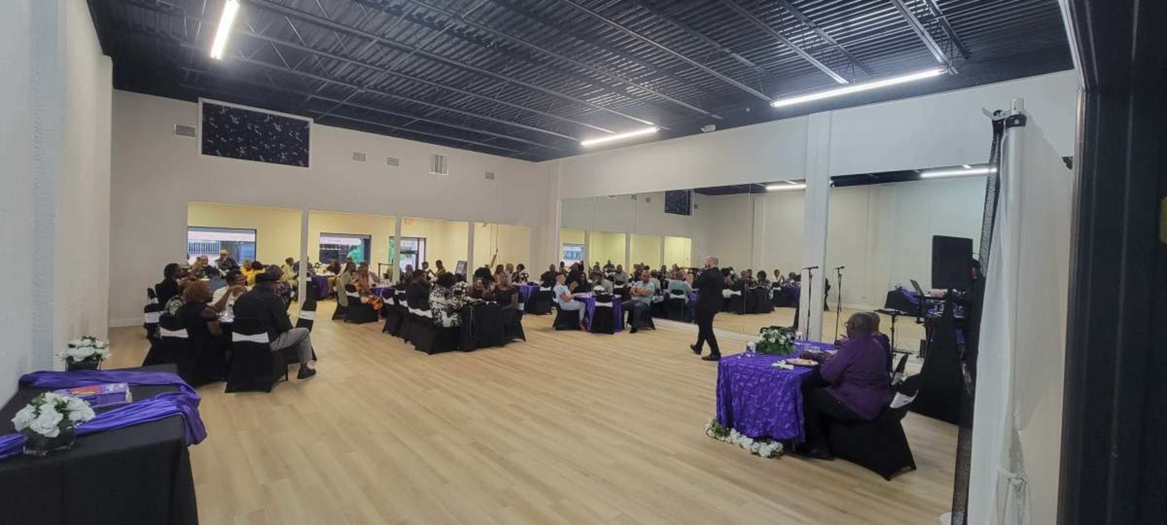 A large event space filled with attendees seated at tables, while a speaker presents in front.