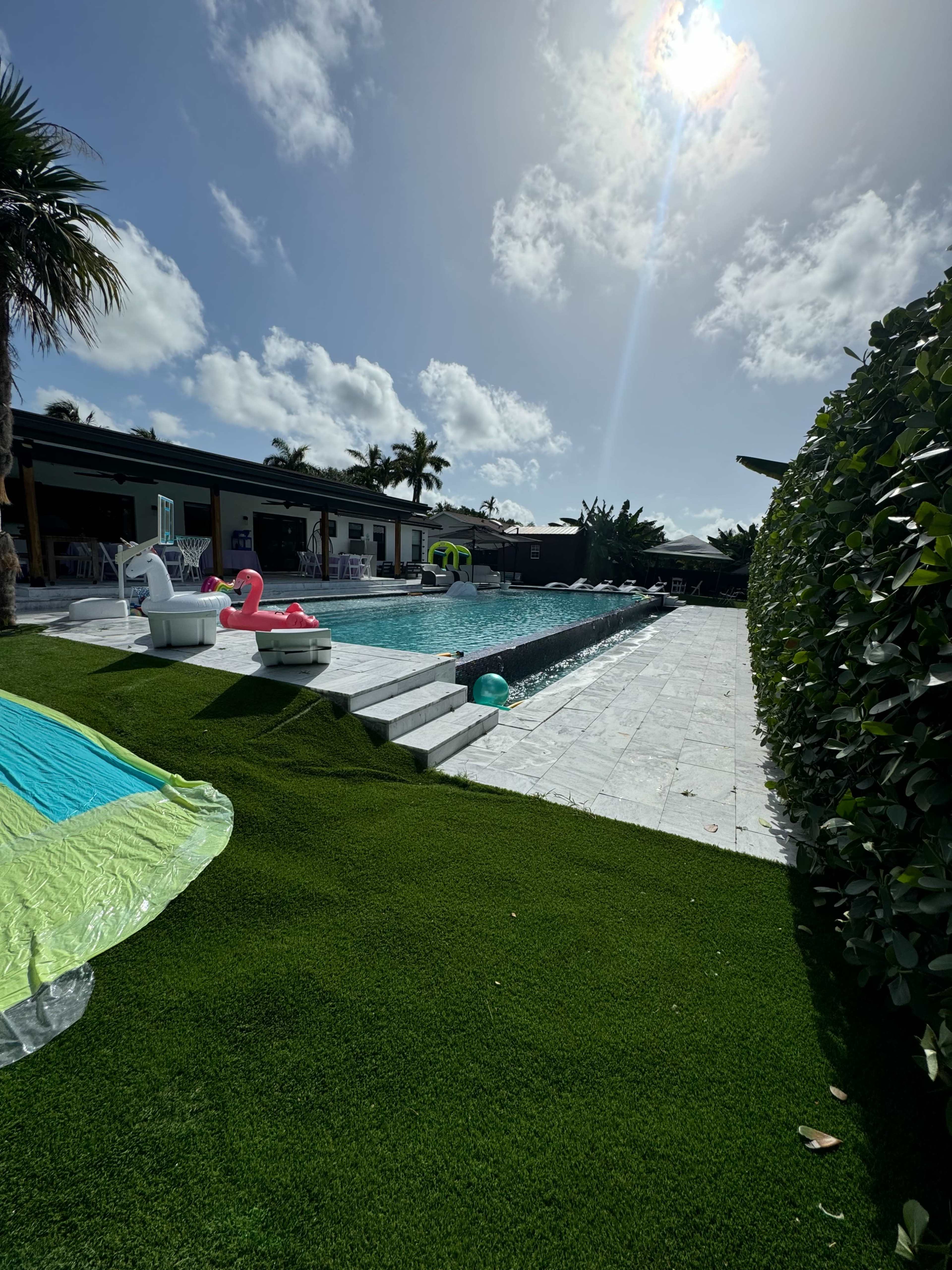 A swimming pool surrounded by greenery, chairs, and colorful pool floats under a bright sky.