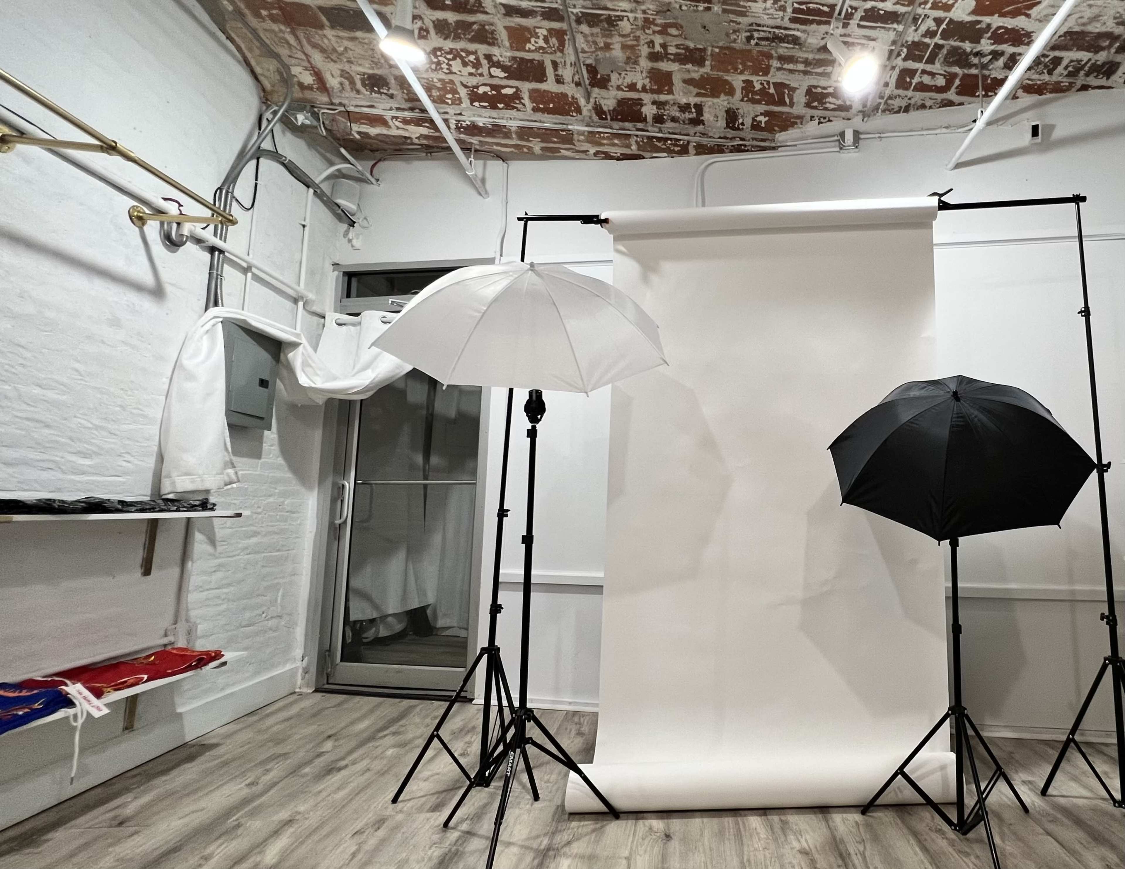 The image shows a photography studio with a white backdrop, two umbrellas (one white and one black) on tripods, and exposed brick walls.
