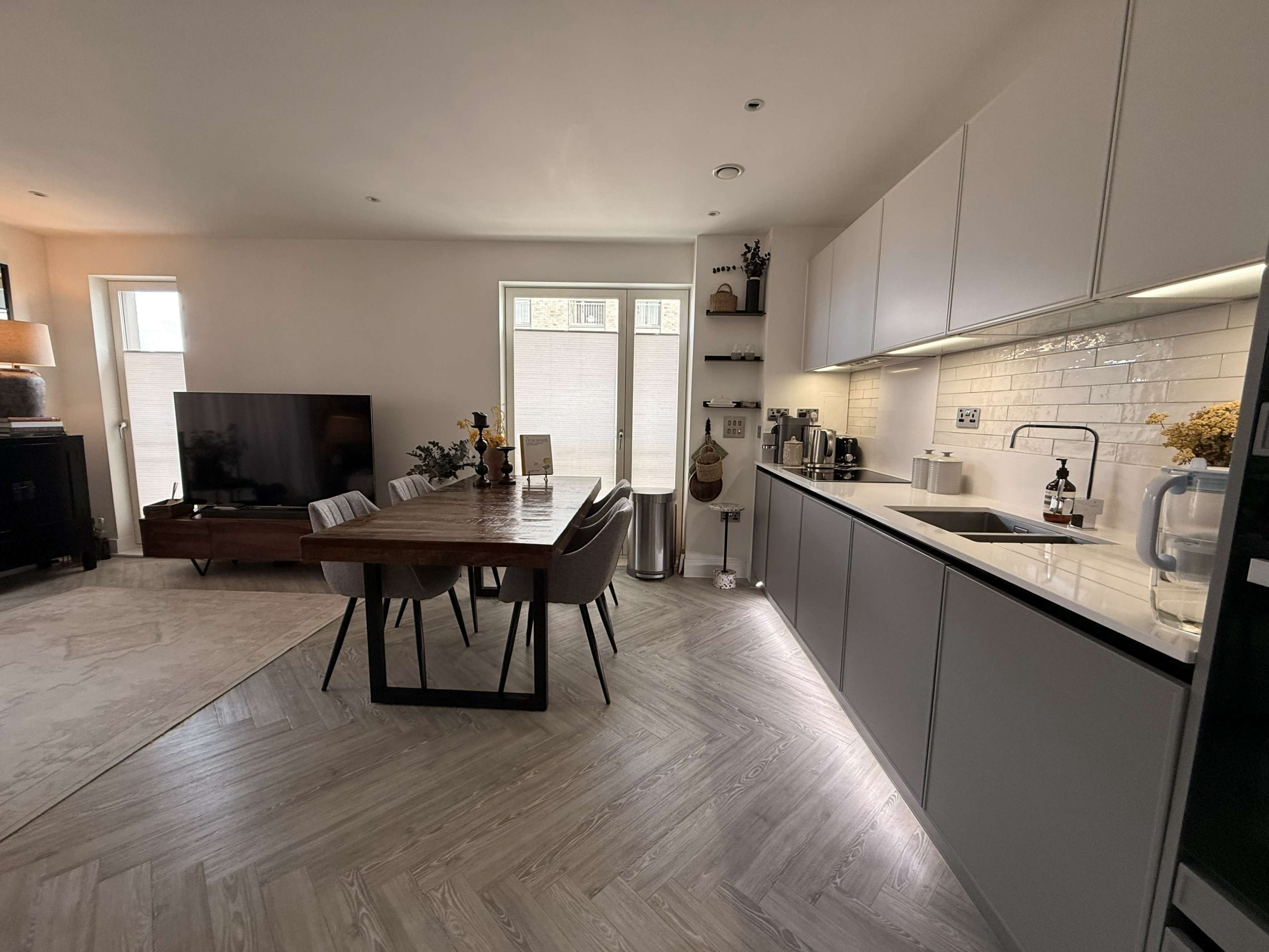 The image shows a modern kitchen with a dining area featuring a wooden table and chairs, along with a sleek countertop and appliances.