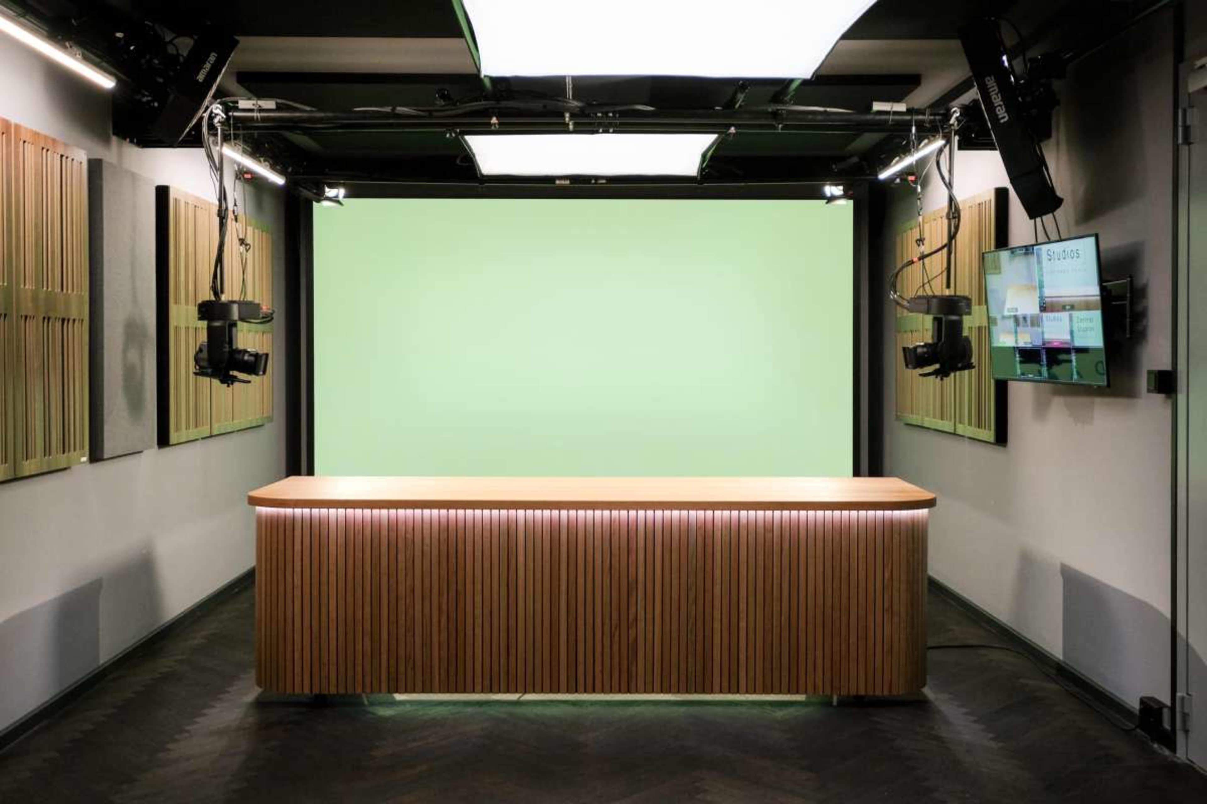 The image shows a modern studio setup featuring a wooden reception desk, green backdrop, and overhead lights, along with camera equipment mounted on the ceiling.