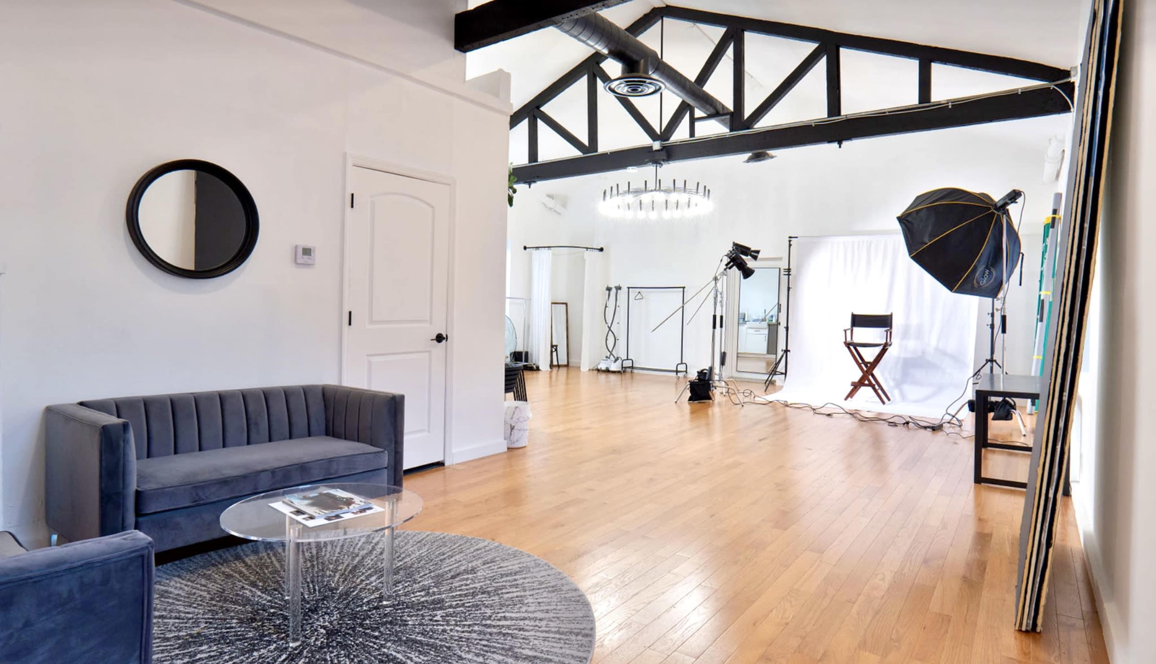 The image shows a spacious studio with a gray sofa, a round glass table, and photography equipment set up against a bright backdrop.