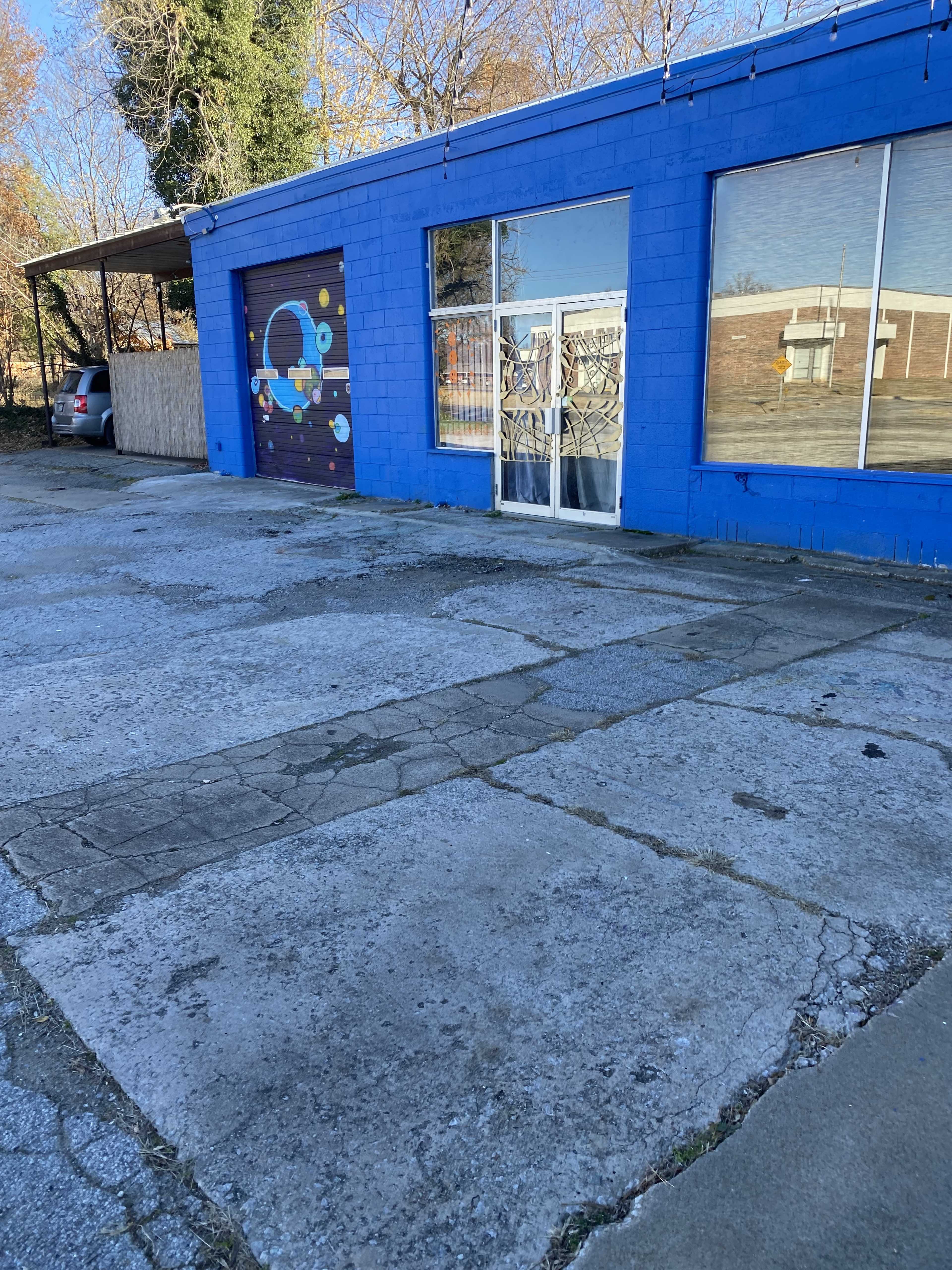 The image shows a blue building with large windows, a garage door painted with colorful designs, and a concrete parking area with visible cracks.