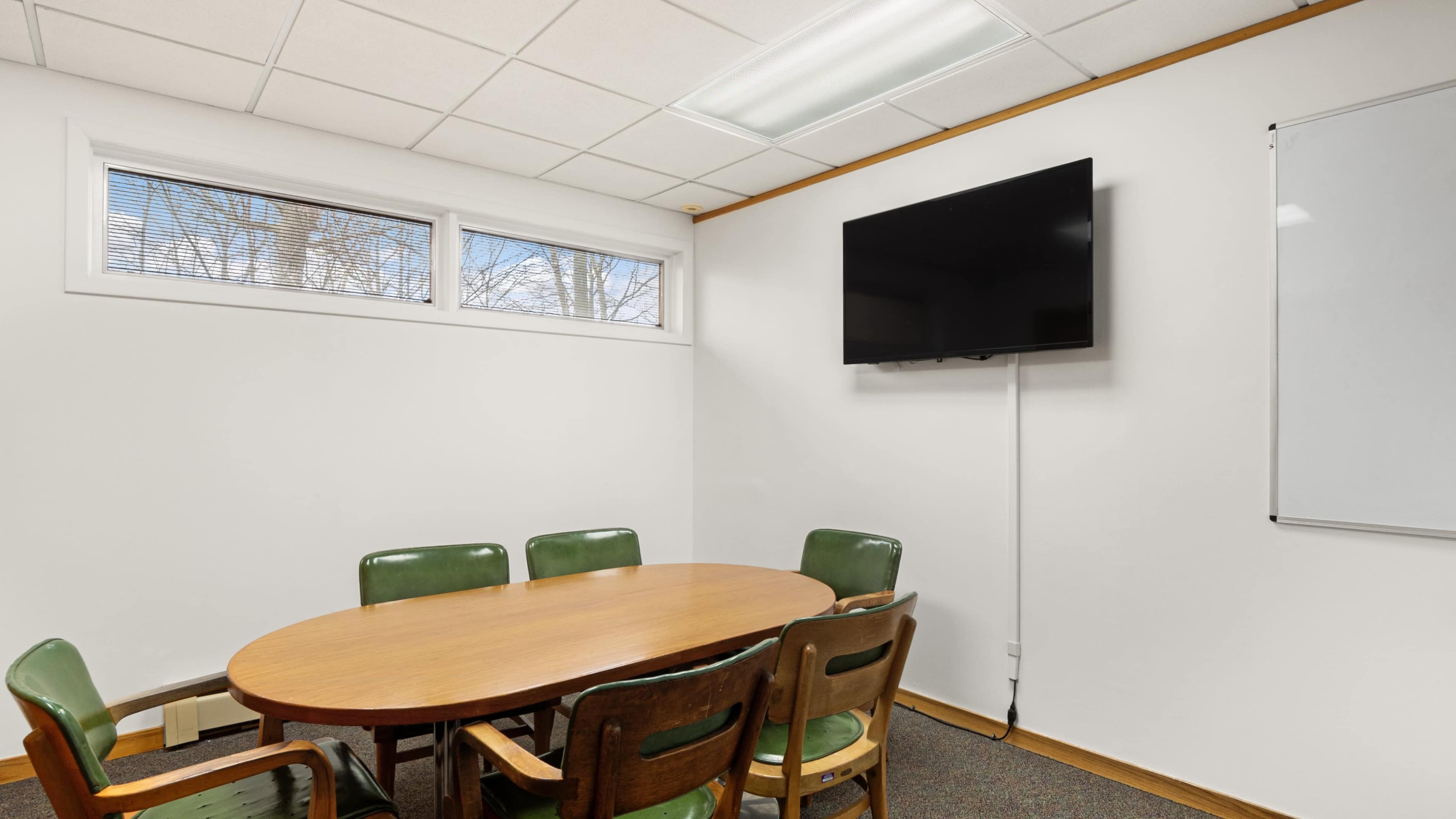 A conference room features a round wooden table surrounded by green and brown chairs, with a large television mounted on the wall and windows providing natural light.