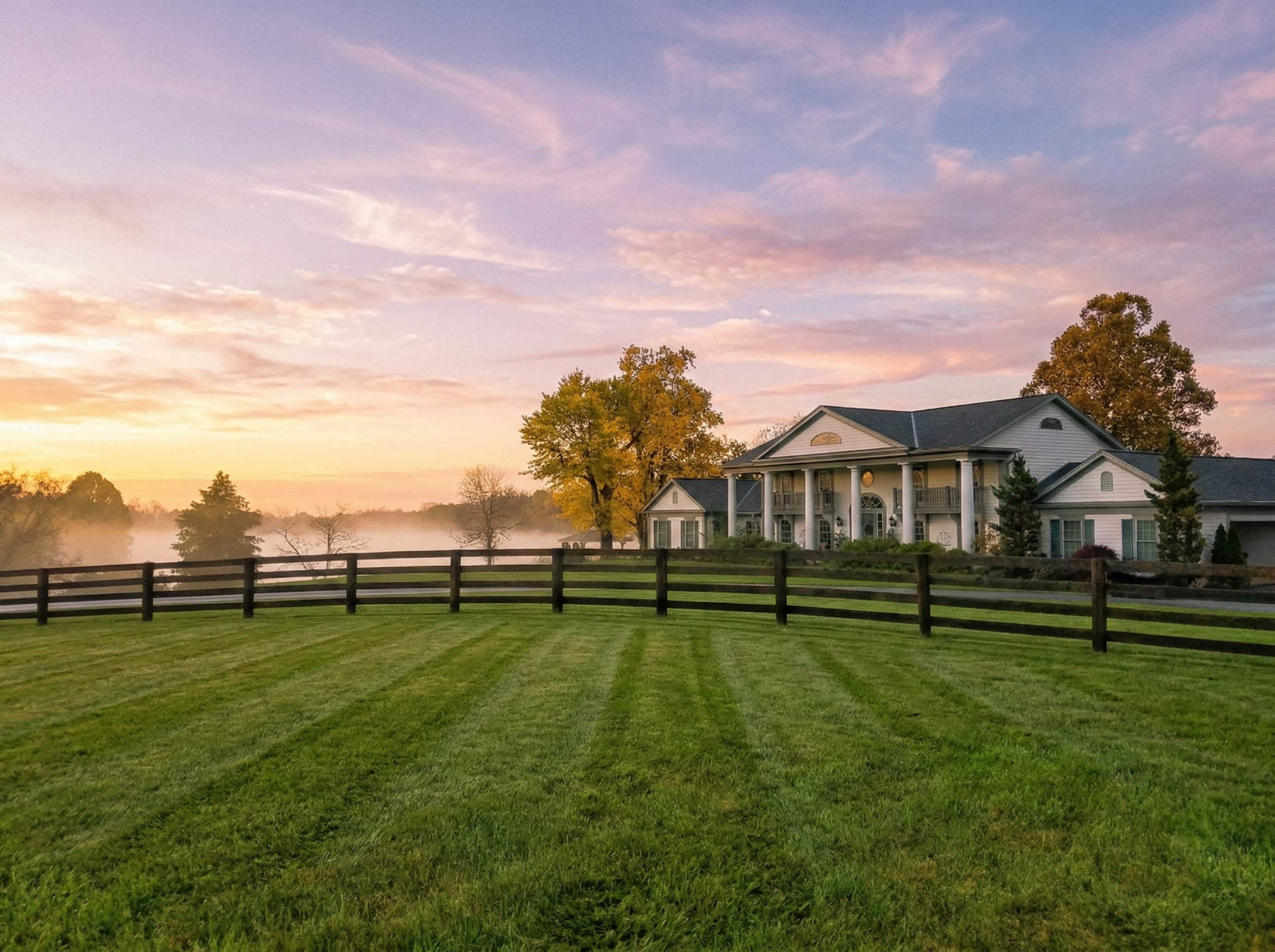 A large, elegant house with a porch stands near a foggy field at sunrise, enclosed by a black wooden fence.