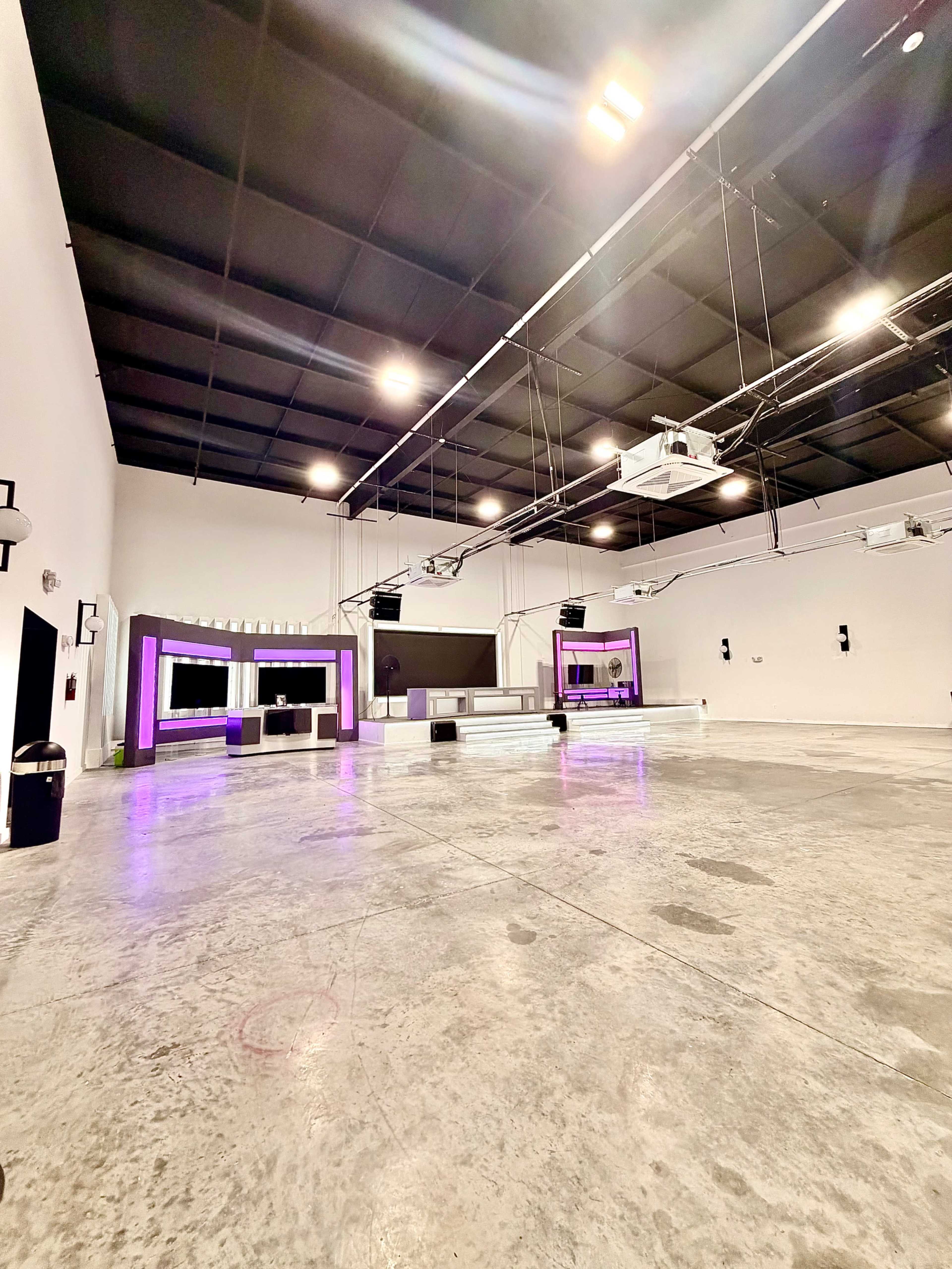 The image shows a spacious, empty studio with polished concrete floors, illuminated by overhead lighting and featuring a purple-lit reception area on one side.
